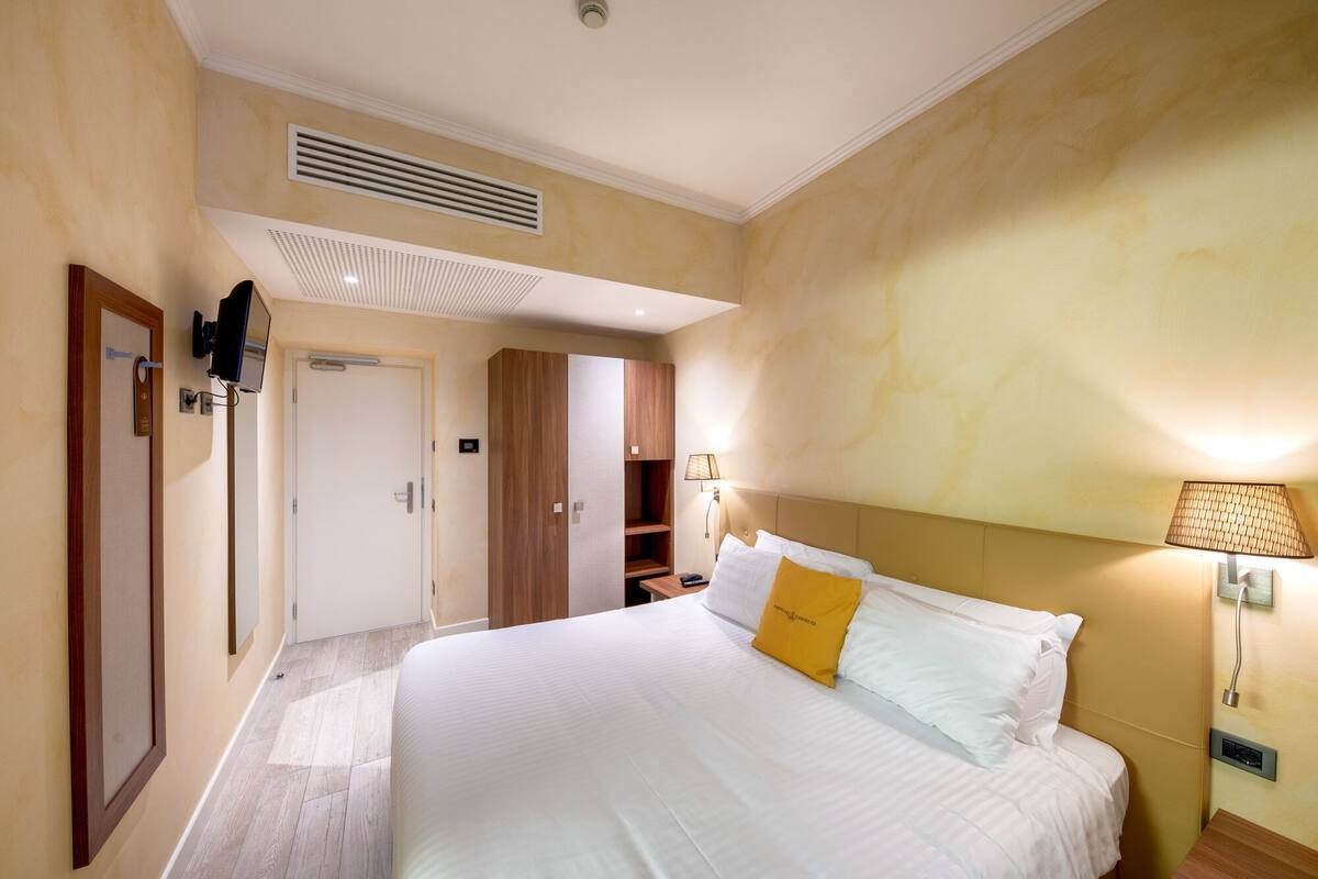 A hotel room with a large bed, a wardrobe, a mirror, a bedside table with a lamp, and a ceiling vent.