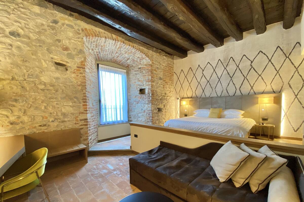 A bedroom with a stone wall, a bed with white bedding, a wooden headboard, and a small table with a lamp.