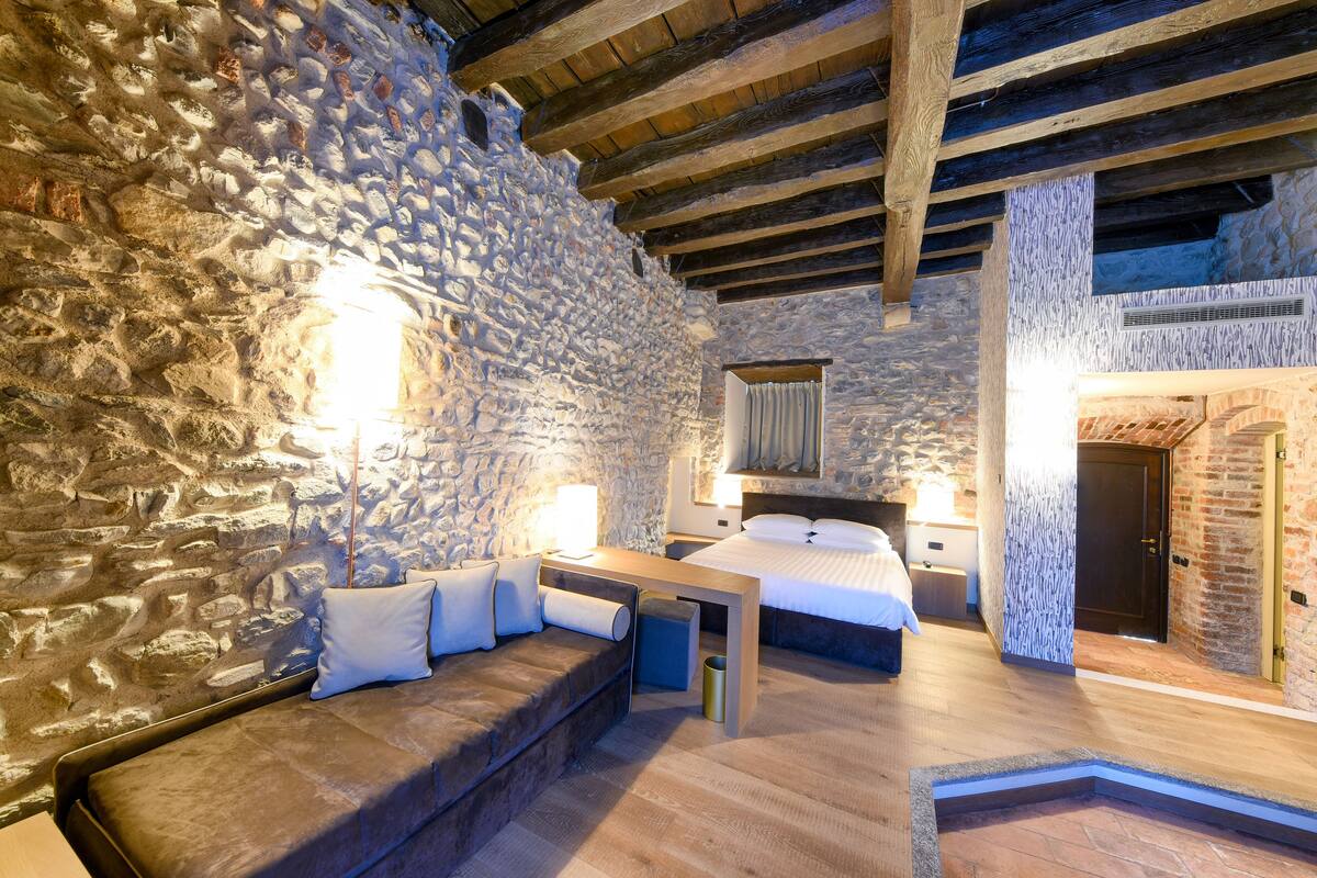 A stone-walled room with a wooden ceiling, a bed, a sofa, and a small table.