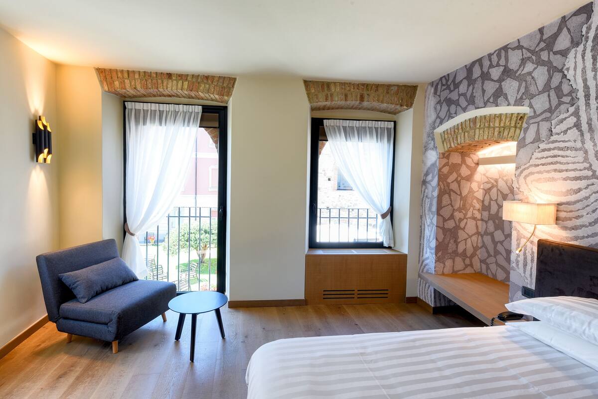 A hotel room with a bed, a sofa, a small table, and two windows with curtains.