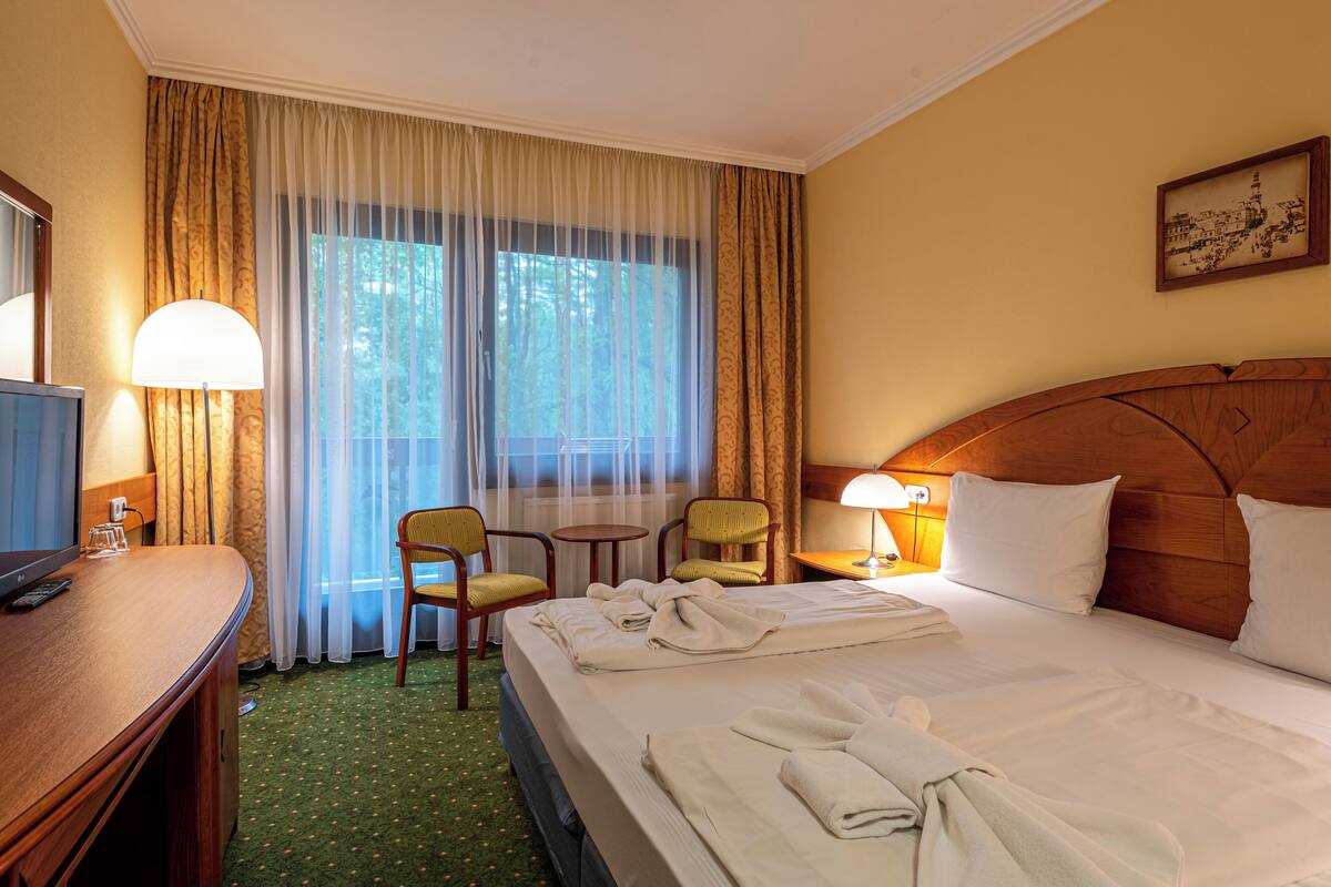 A hotel room with a bed, two chairs, a desk with a TV, and a window with curtains.