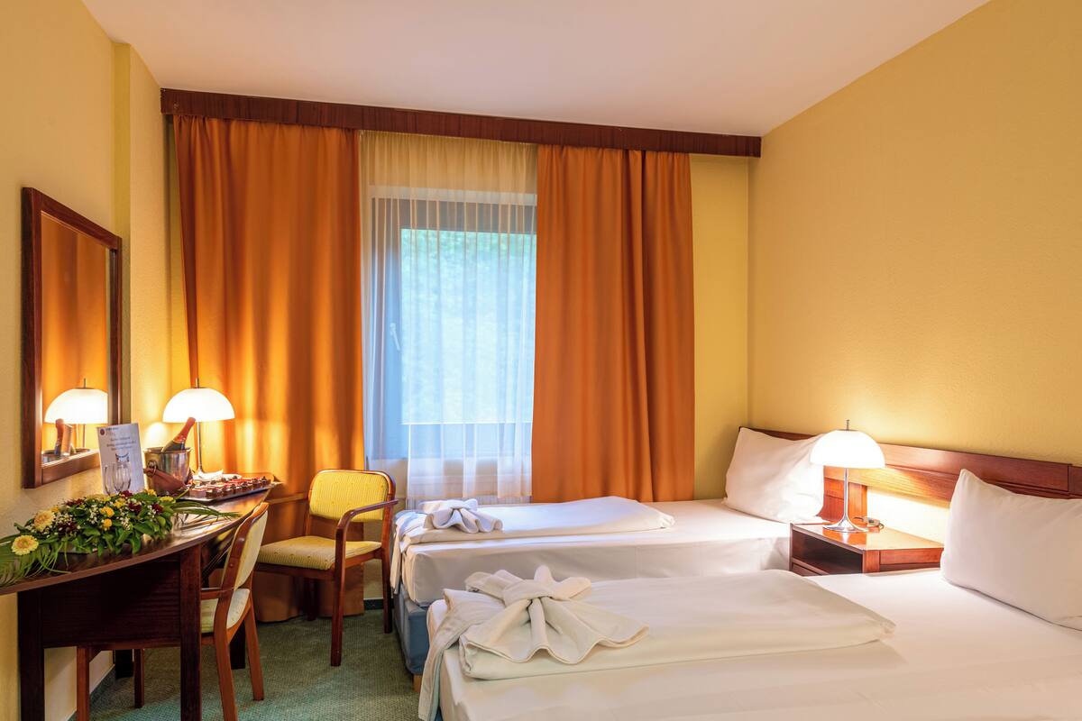 A hotel room with two beds, a desk with a lamp, a chair, and a window with curtains.
