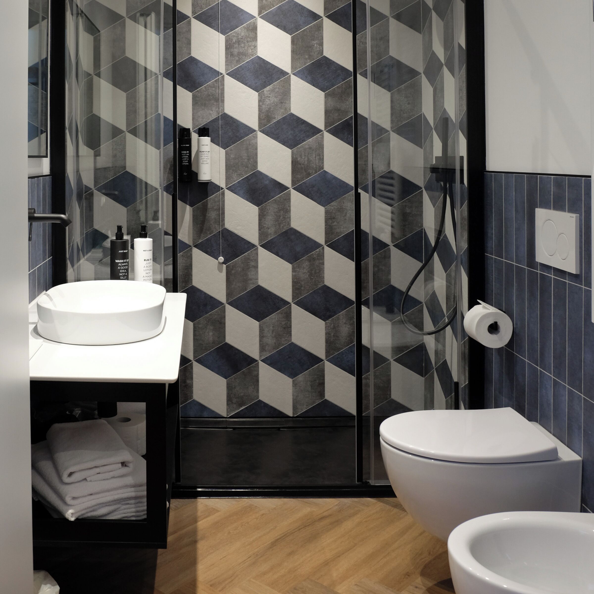 A modern bathroom with a geometric tile backsplash, a white sink, a toilet, and a glass shower enclosure.