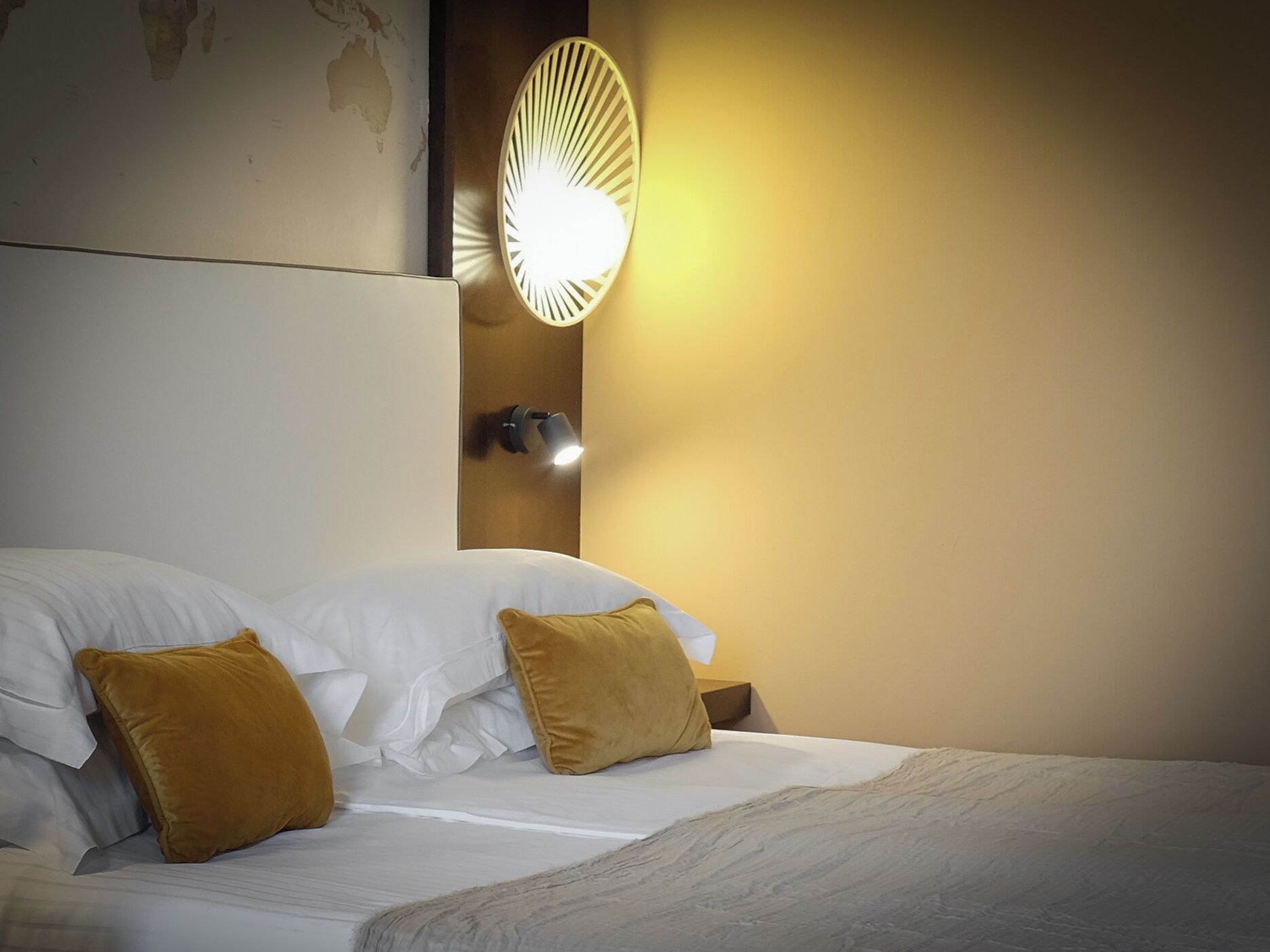 A neatly made bed with white linens and two mustard-colored pillows, a wall-mounted light fixture, and a partial view of a world map on the wall.