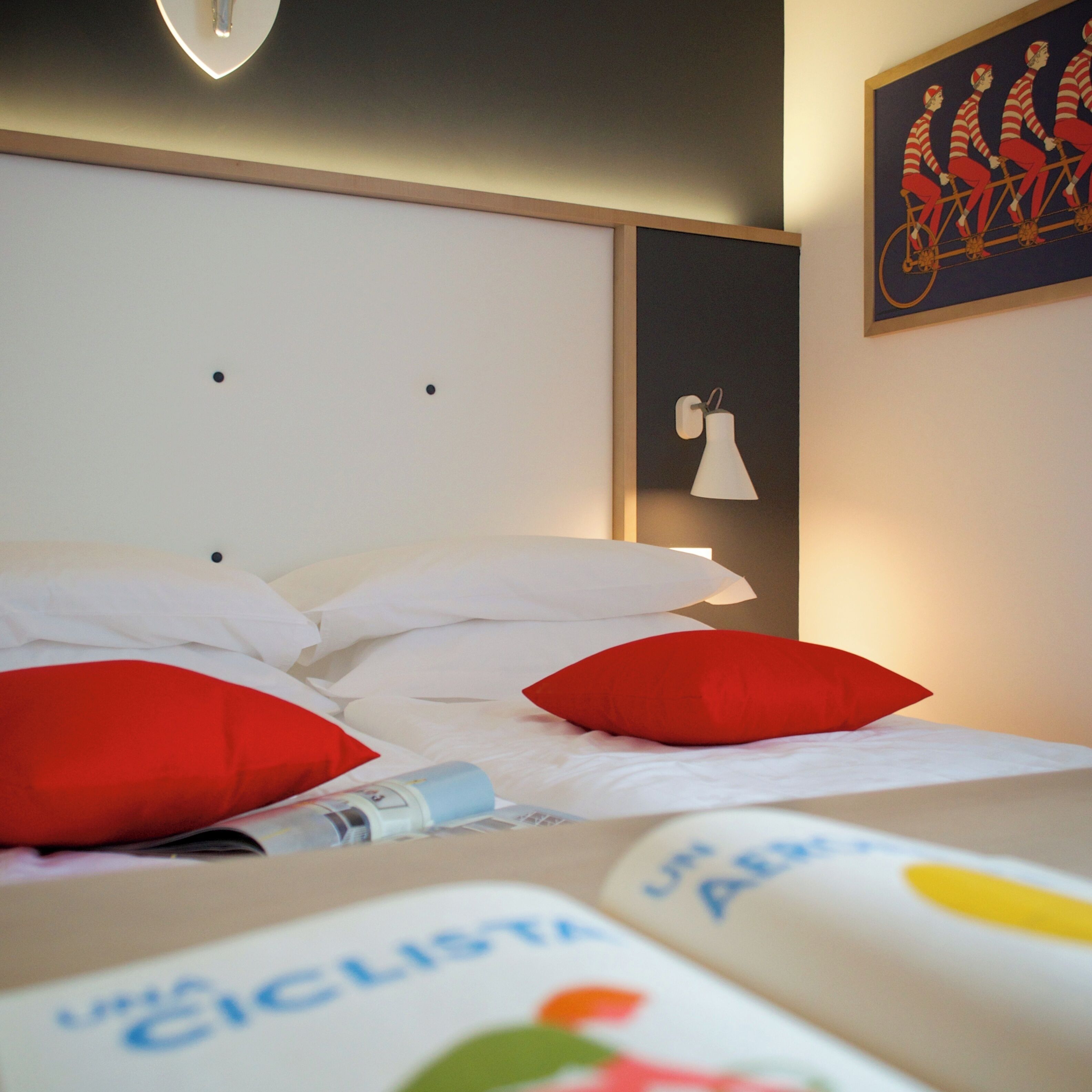 A neatly made bed with white and red pillows, a headboard with a light fixture, and a wall-mounted lamp.