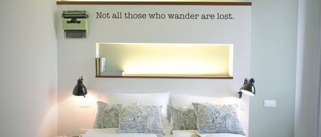 Un dormitorio ordenado con cama, almohadas, mesita de noche, lámpara y una estantería de pared con una cita enmarcada.