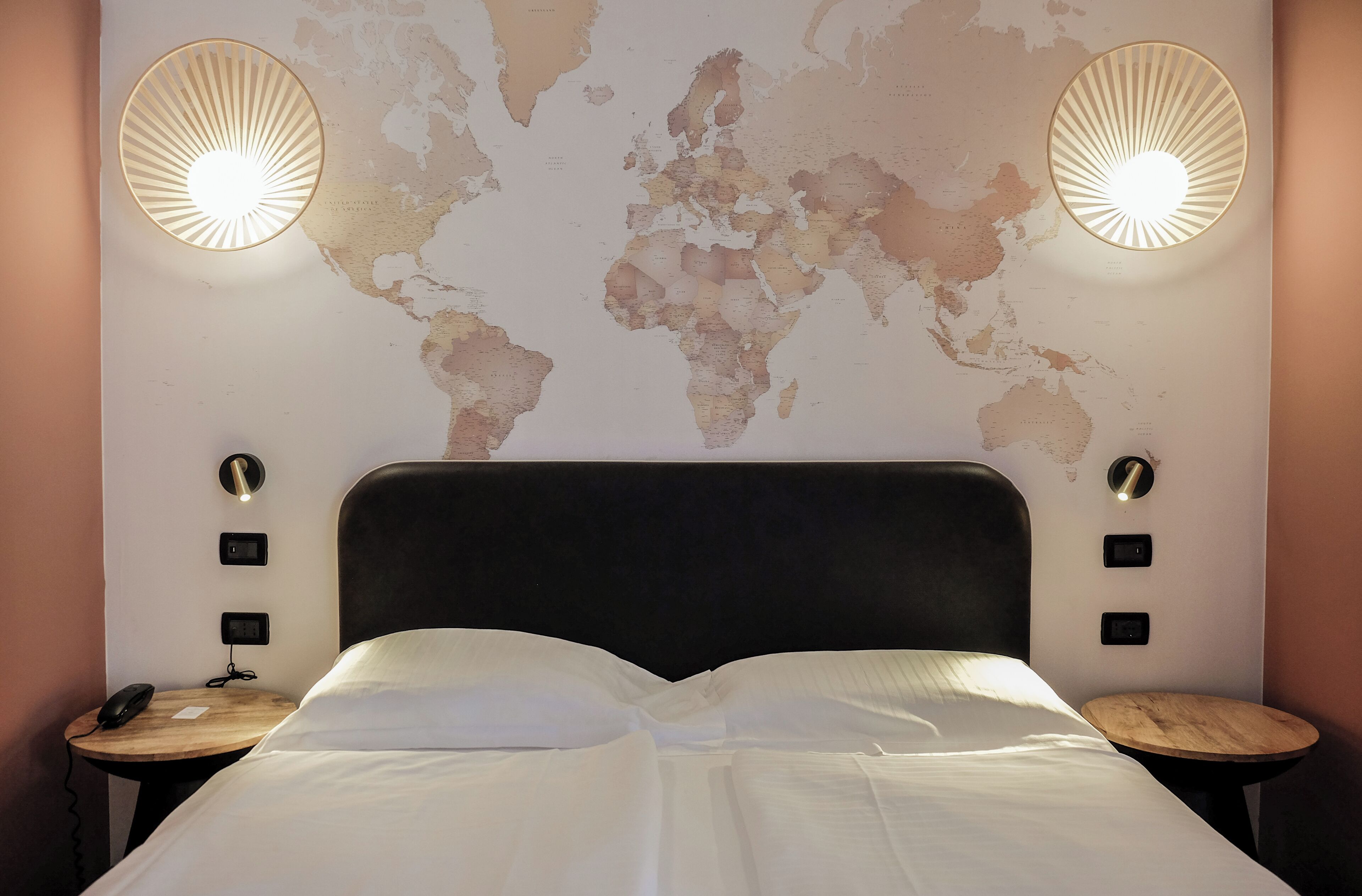 Une chambre d’hôtel avec une carte du monde accrochée au mur, une tête de lit noire et deux tables de chevet avec des lampes.
