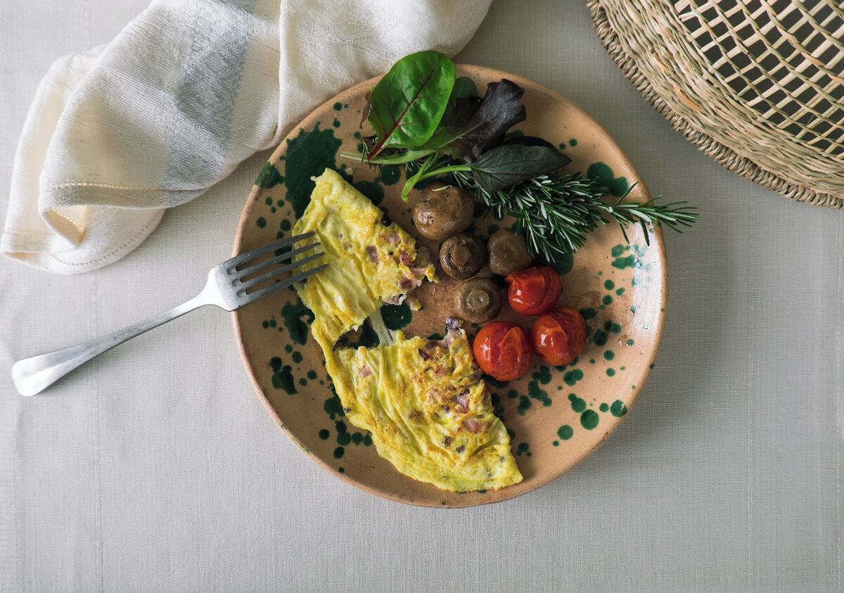 A plate with an omelette, mushrooms, tomatoes, and greens on a textured tablecloth.