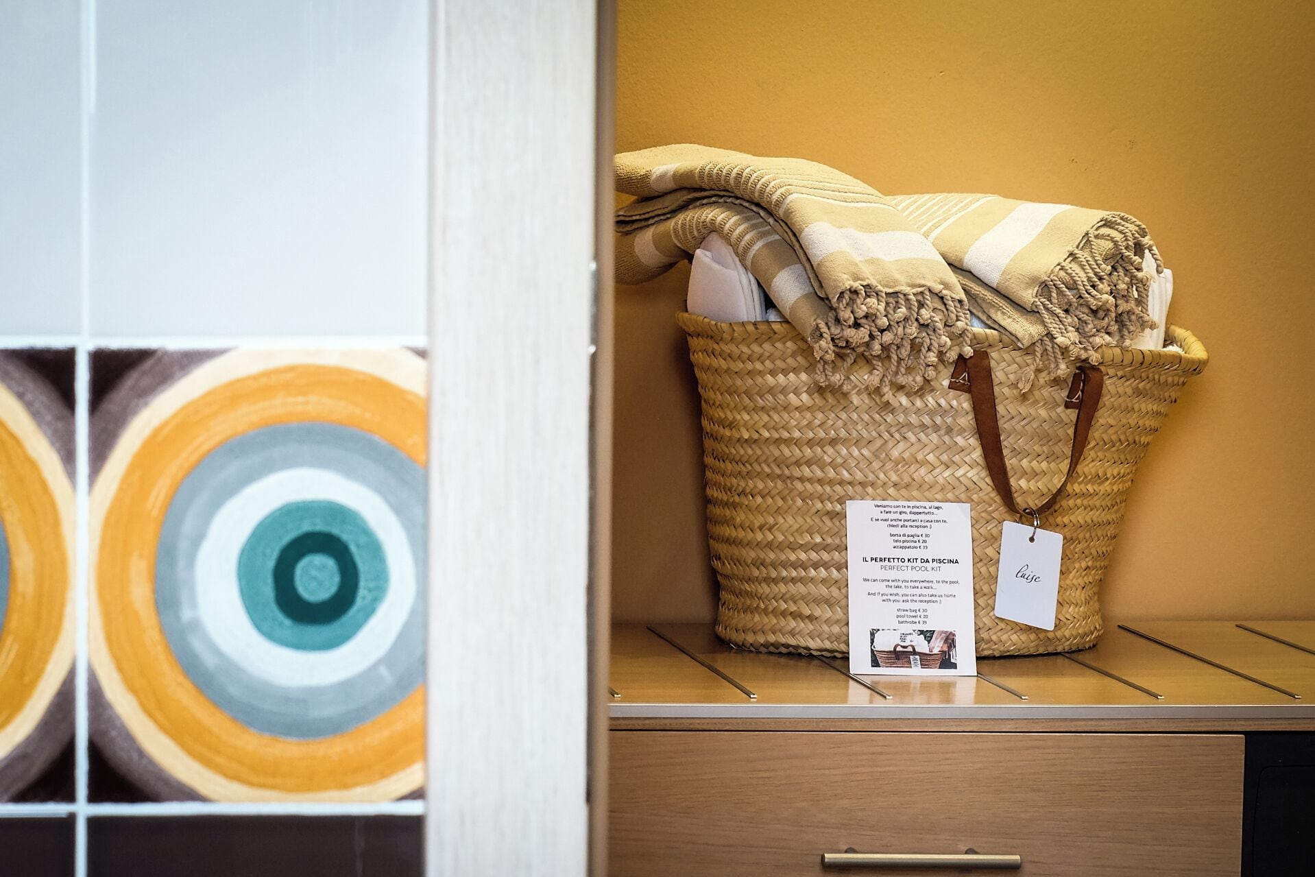 A woven basket with a tag, placed on a wooden shelf against a yellow wall.
