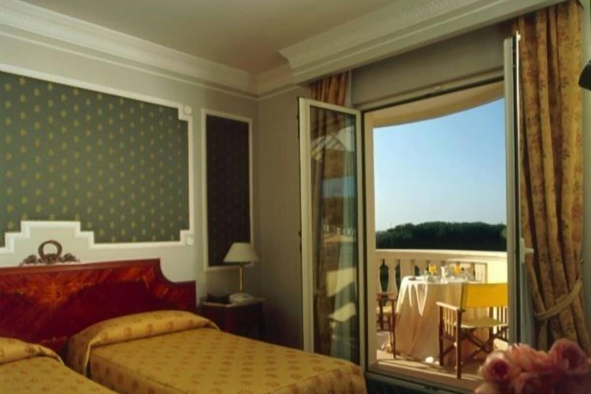 Habitación de hotel con dos camas, ventana con vistas y balcón con mesa y sillas.