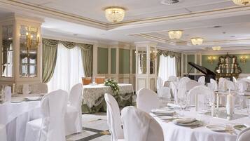 A formal dining room with white tablecloths, white chairs, chandeliers, and a grand piano.