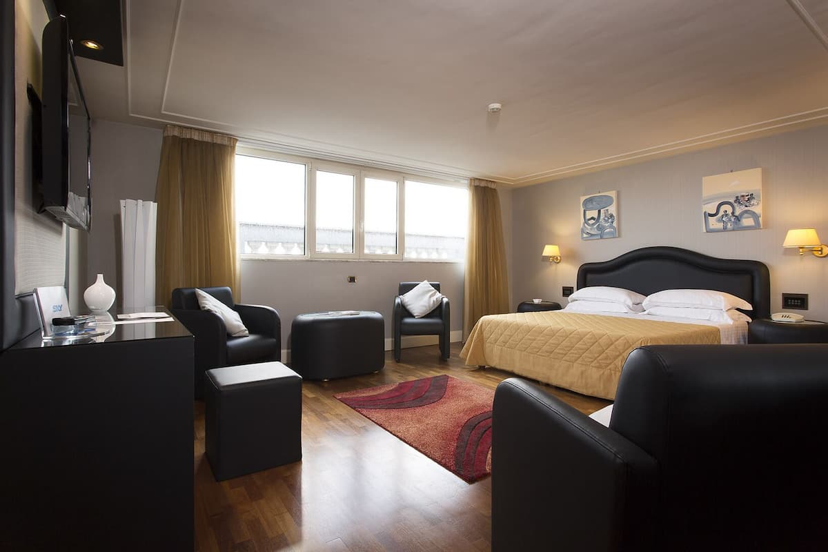 Habitación de hotel con cama, dos sillones, escritorio, televisor y ventana con cortinas.