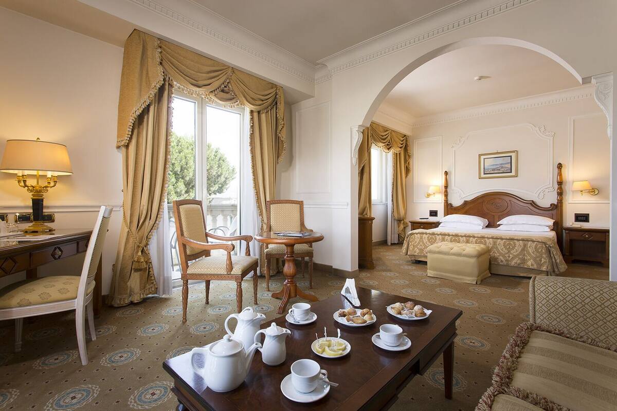 Habitación de hotel con zona de comedor, cama con cabecera y vistas al exterior a través de amplias ventanas.
