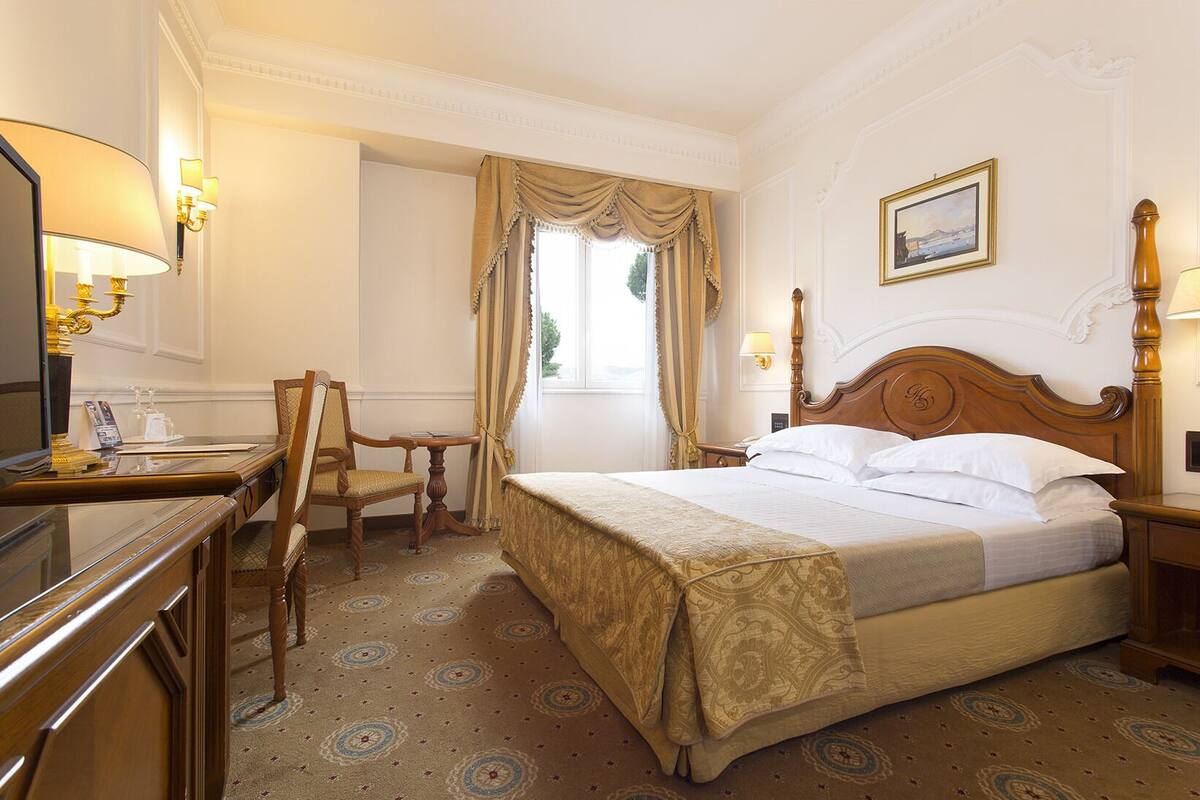 Habitación de hotel con una cama grande, un escritorio con silla, televisión y ventana con cortinas.