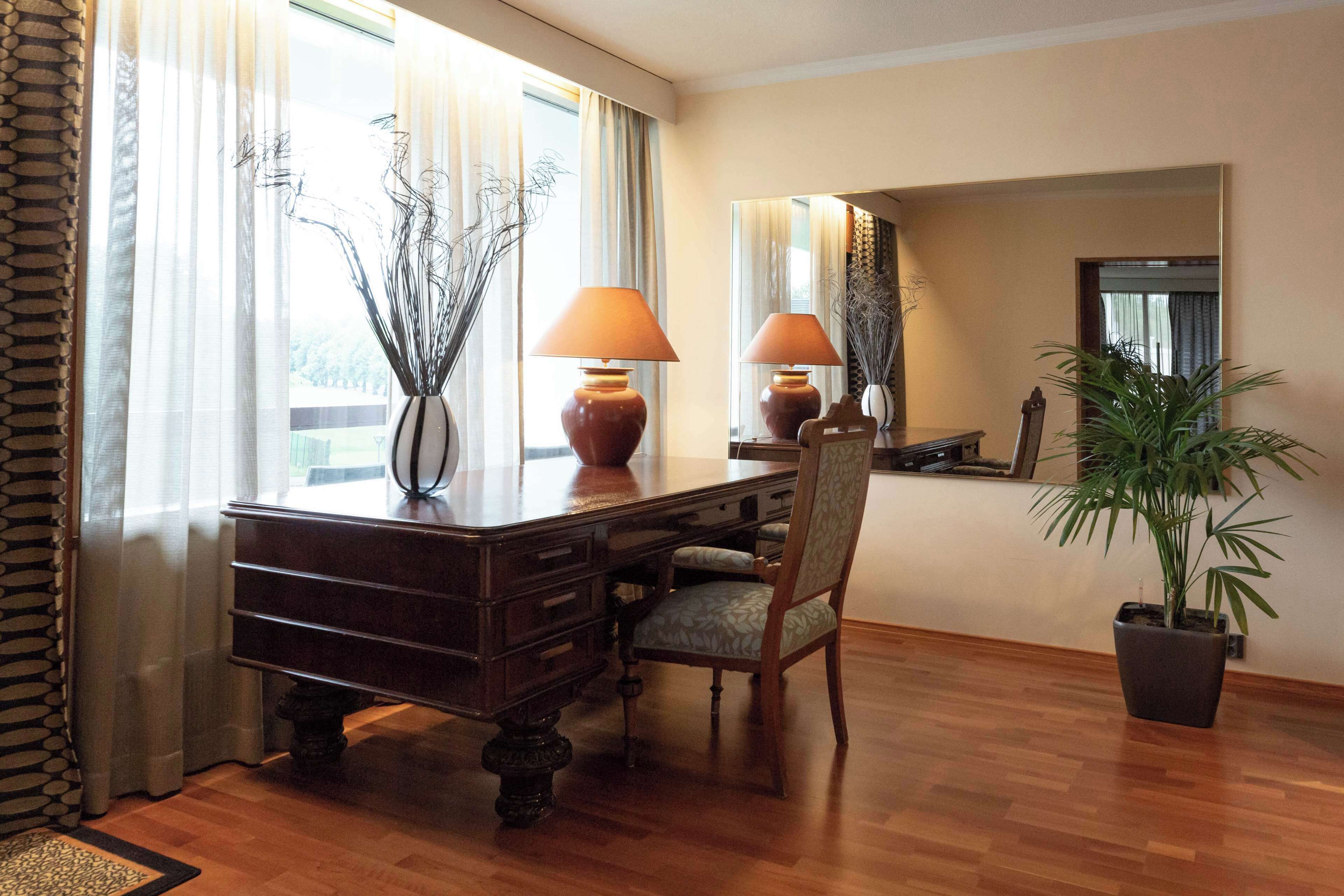 Un escritorio de madera con una lámpara, una silla y una planta en maceta en una habitación con piso de madera y un ventanal con cortinas.