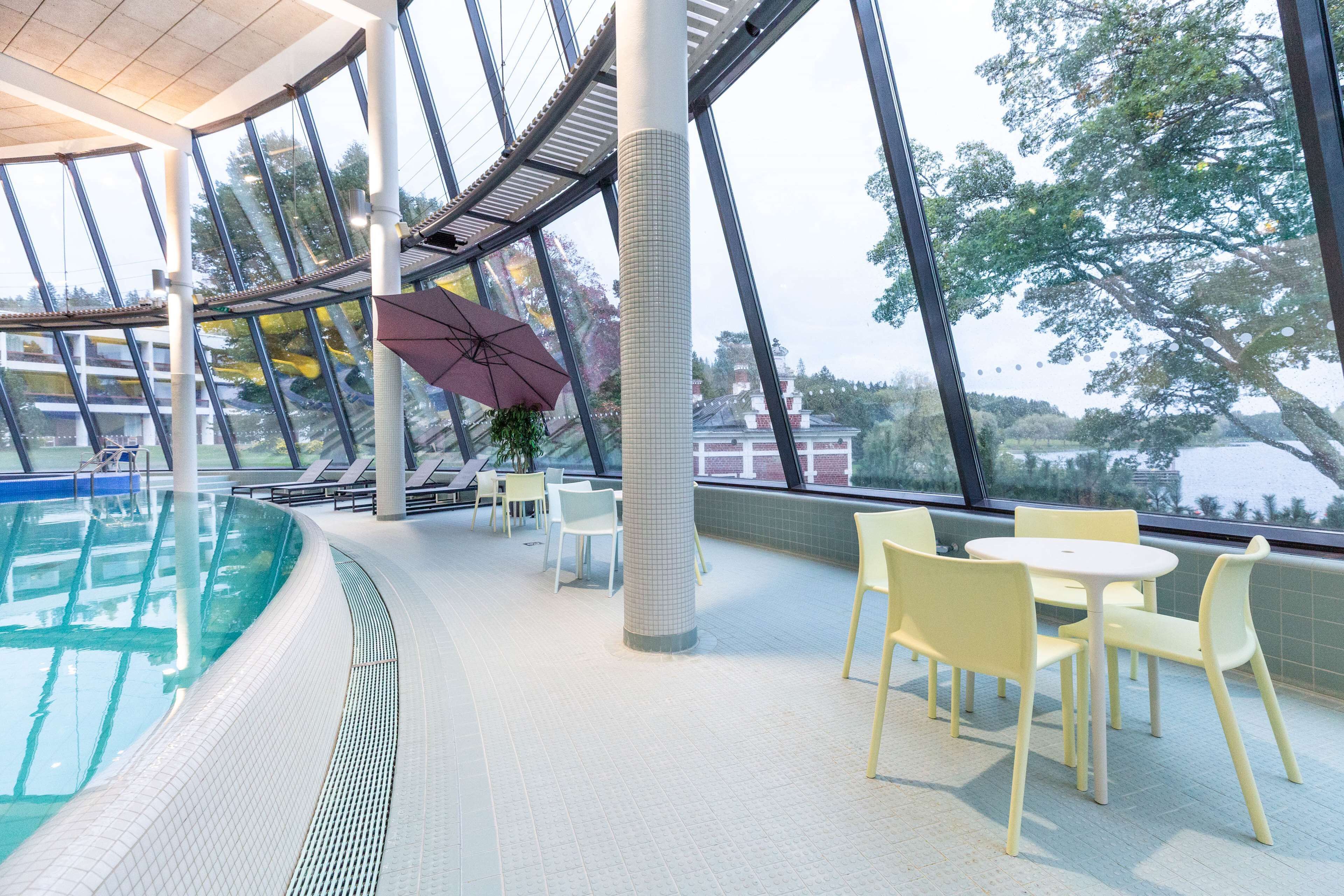 An indoor swimming pool with clear glass walls, yellow chairs, and a table with a red umbrella.