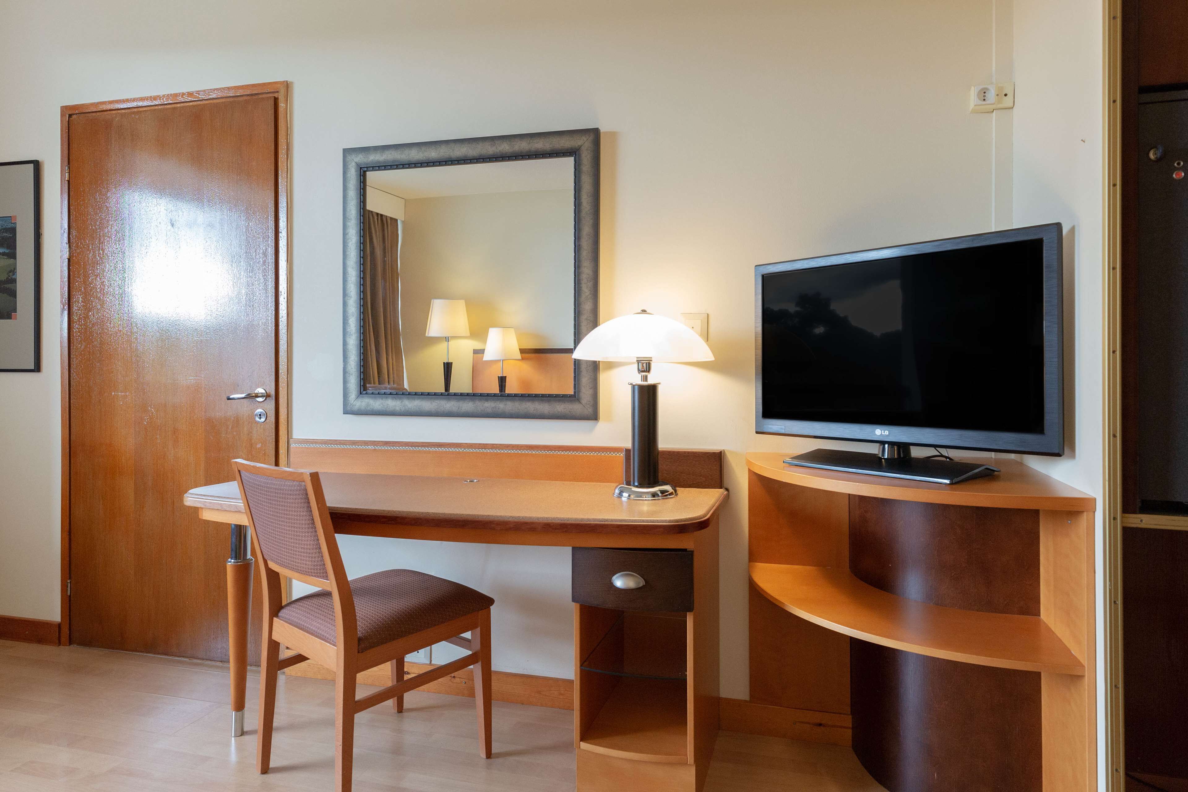A hotel room with a wooden desk, a chair, a television, a lamp, and a mirror.