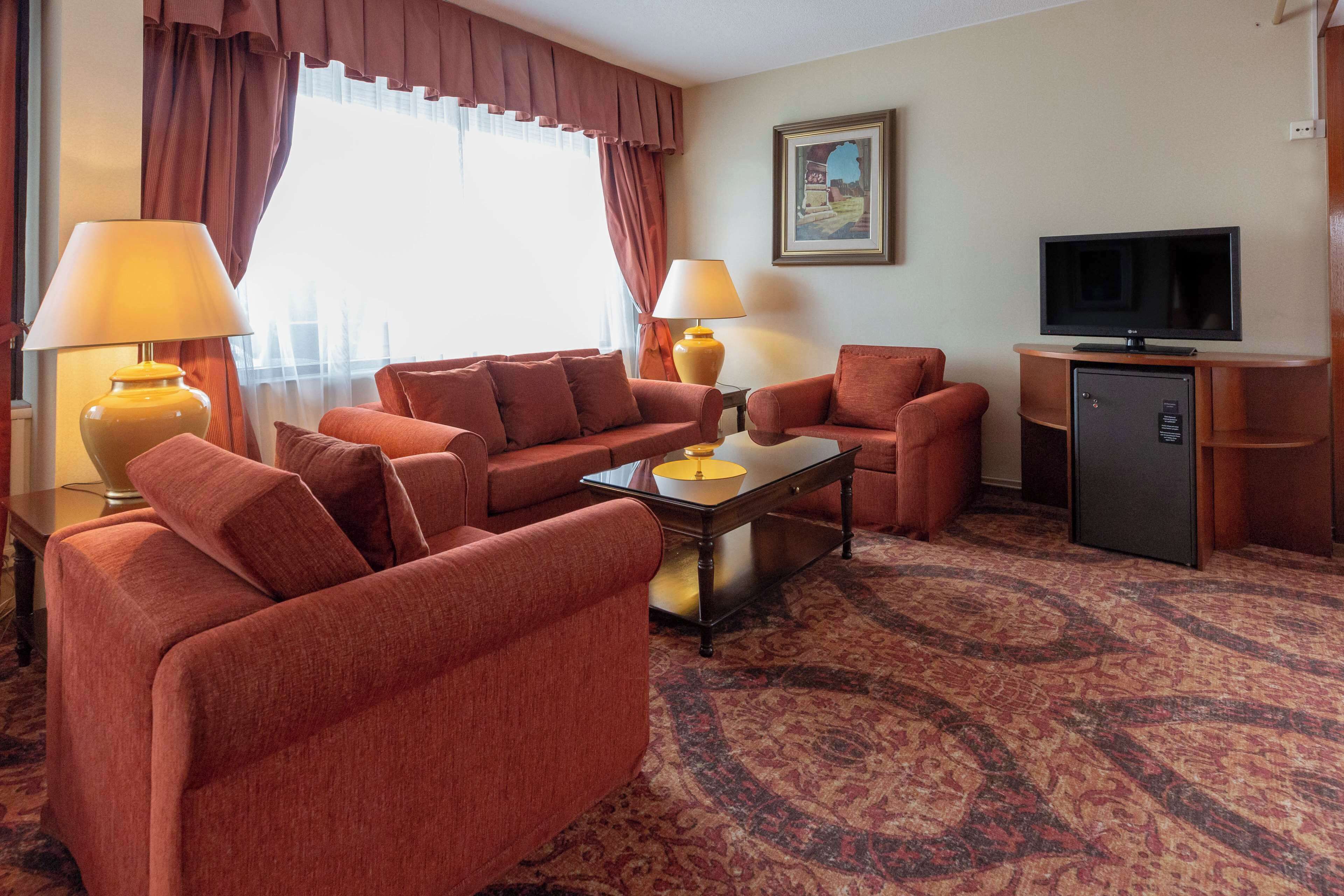 A hotel room with a sofa set, two lamps, a television, and a refrigerator.