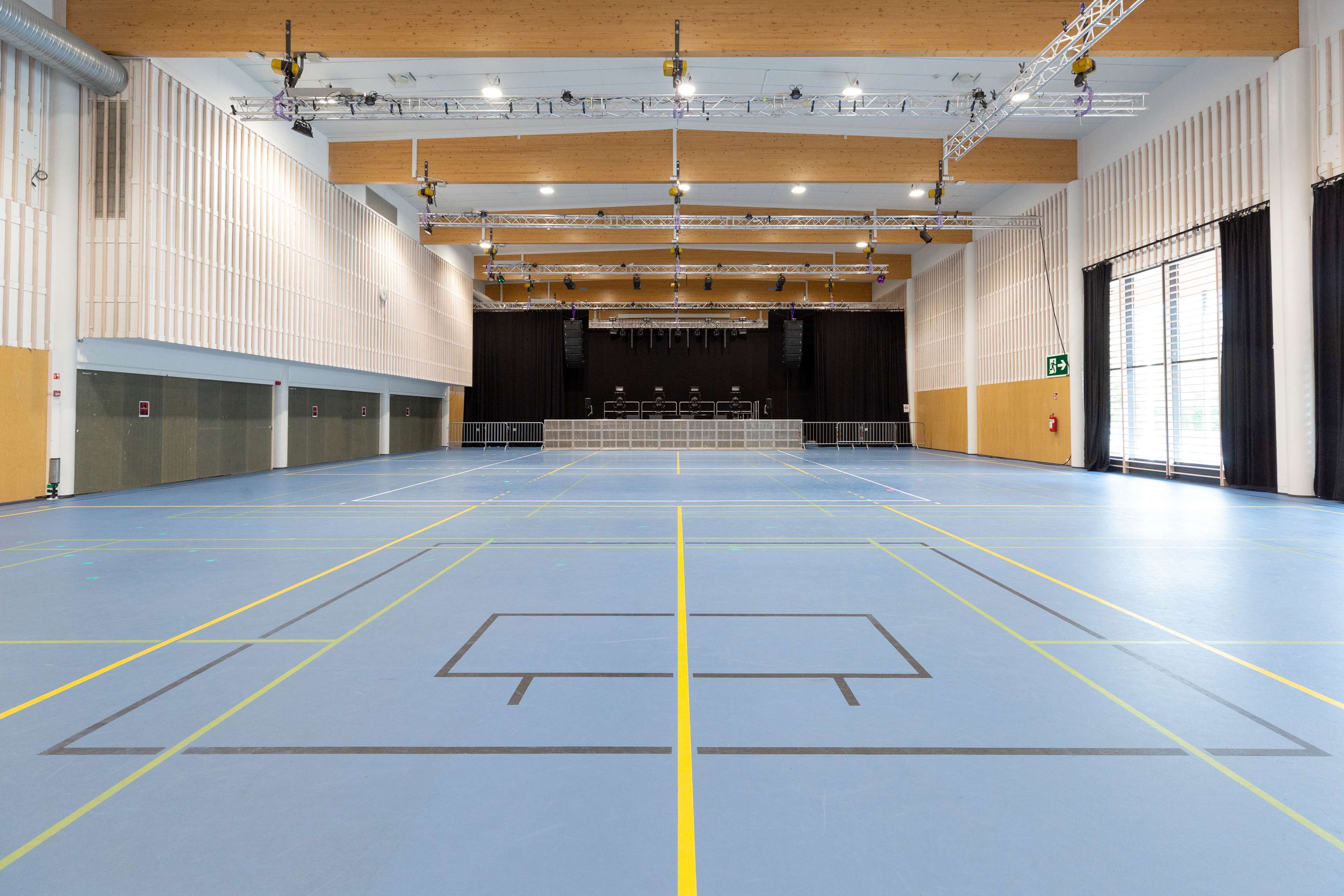 An indoor sports hall with a blue floor marked for sports, a stage with lighting, and a large window on the right.