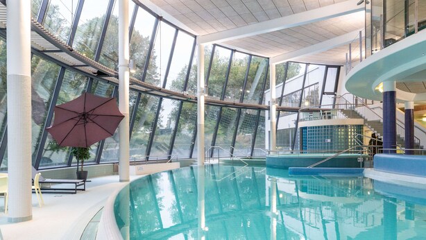 A large indoor swimming pool with clear glass walls, a sun umbrella, and a staircase leading to an upper level. - Scandic Aulanko (Hameenlinna)