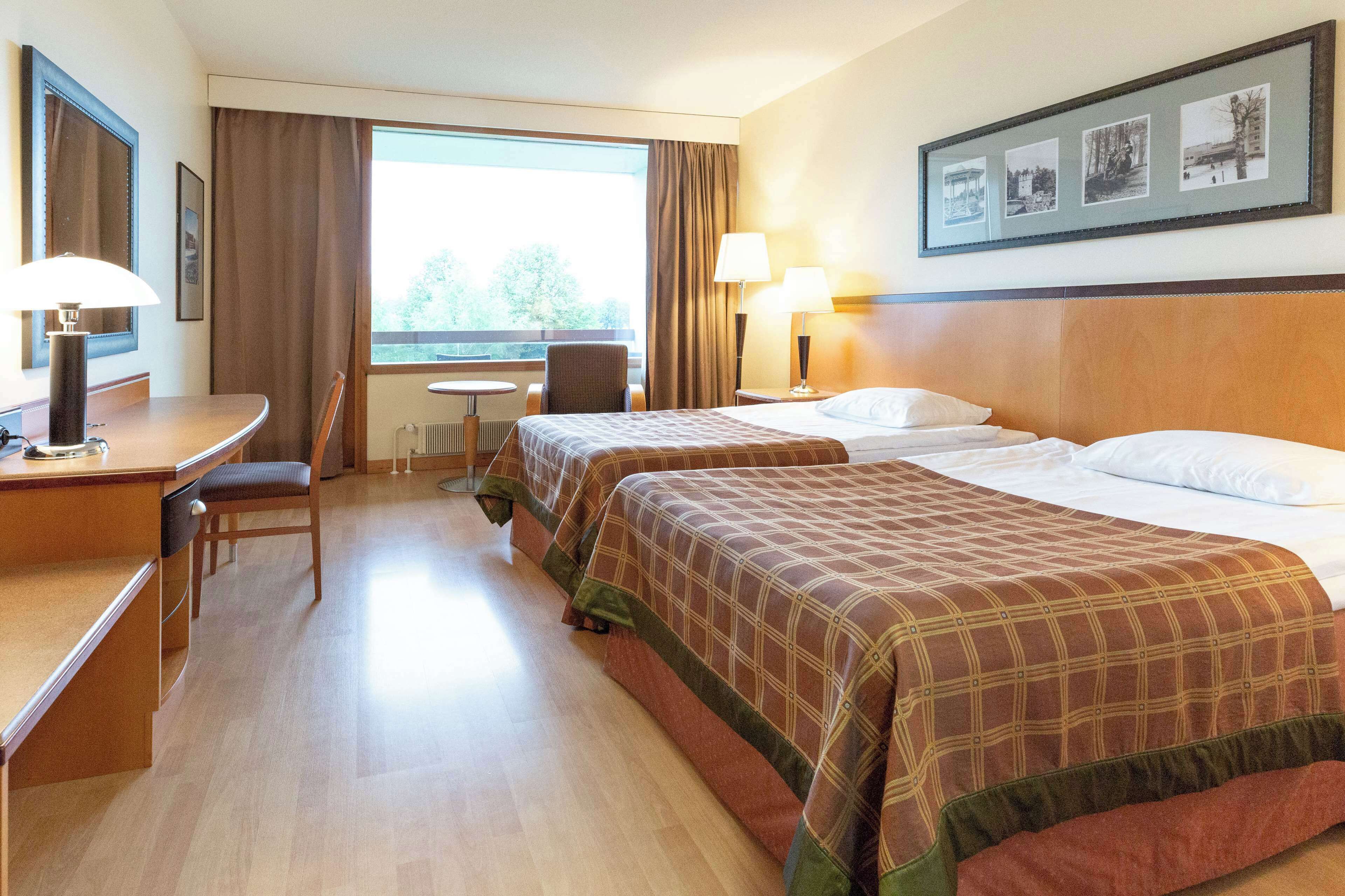 A hotel room with two beds, a desk, a chair, a window with curtains, and a framed picture on the wall.