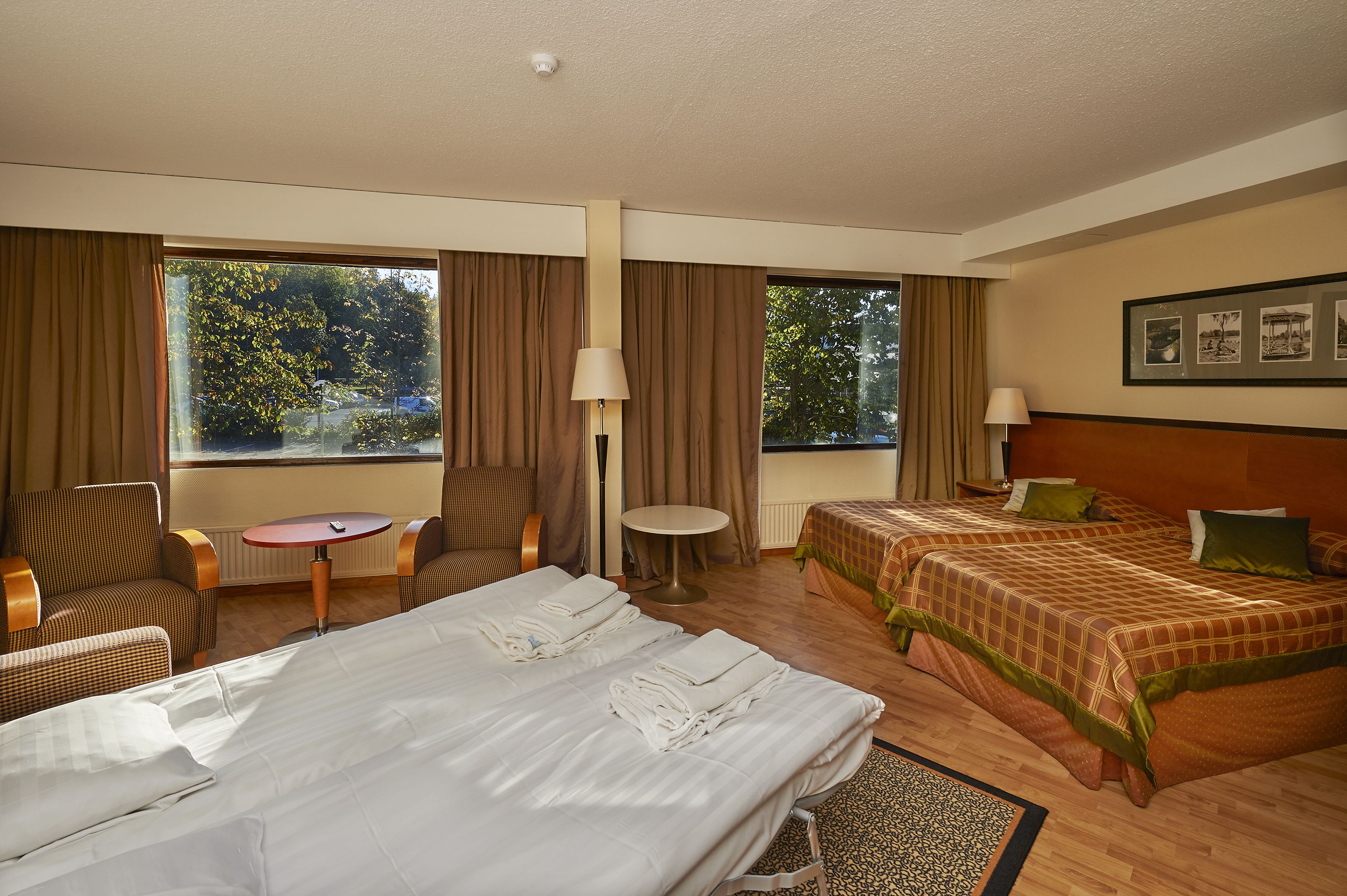 A hotel room with two beds, a sitting area with a table and chairs, and large windows with curtains.