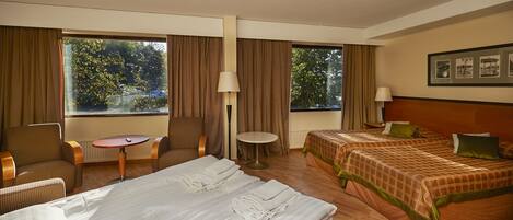 A hotel room with two beds, a sitting area with a table and chairs, and large windows with curtains.