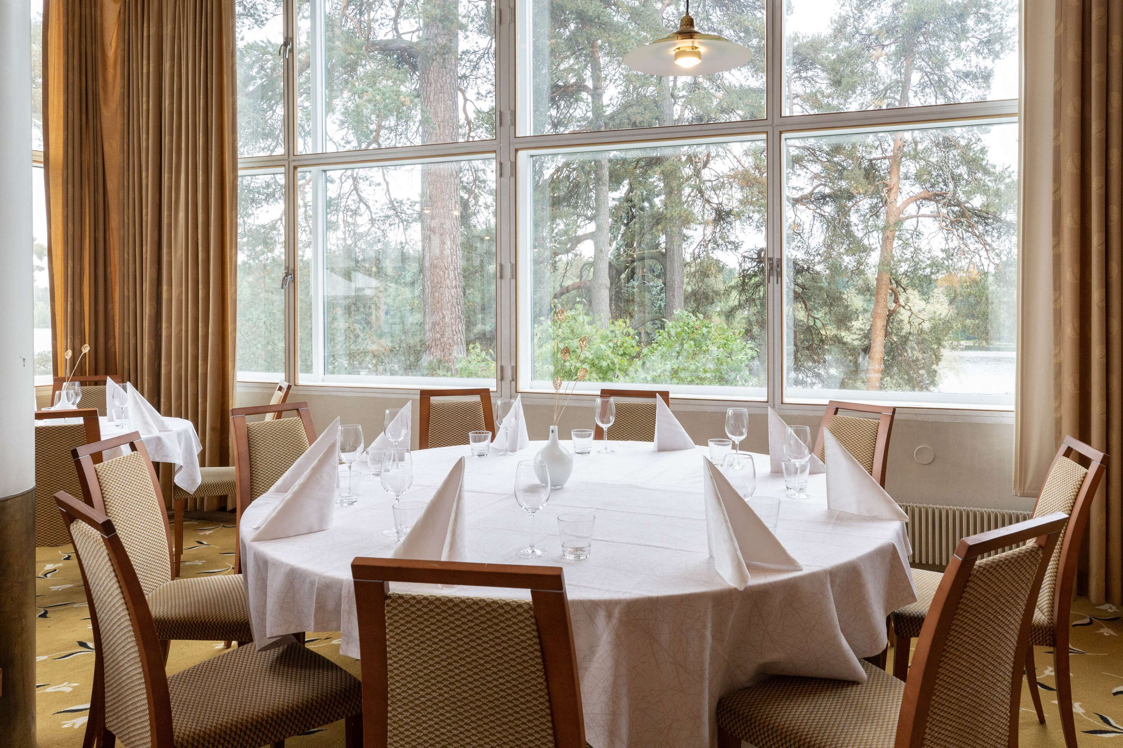 Un comedor con una mesa redonda dispuesta para seis personas, rodeada de sillas de madera con asientos acolchados. Amplias ventanas ofrecen vistas a árboles y una masa de agua.