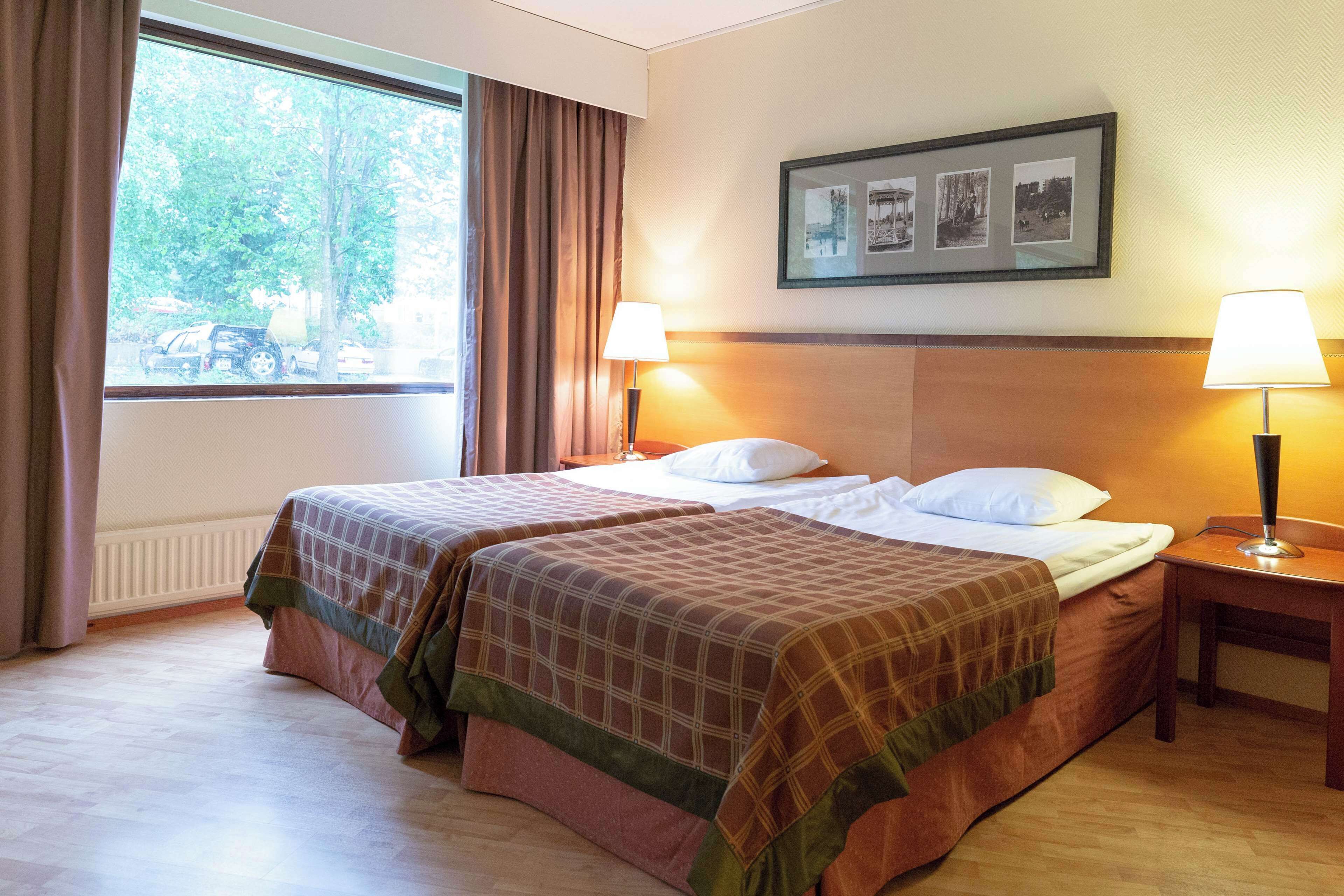 A hotel room with two beds, a window with curtains, a framed picture on the wall, and two bedside lamps.