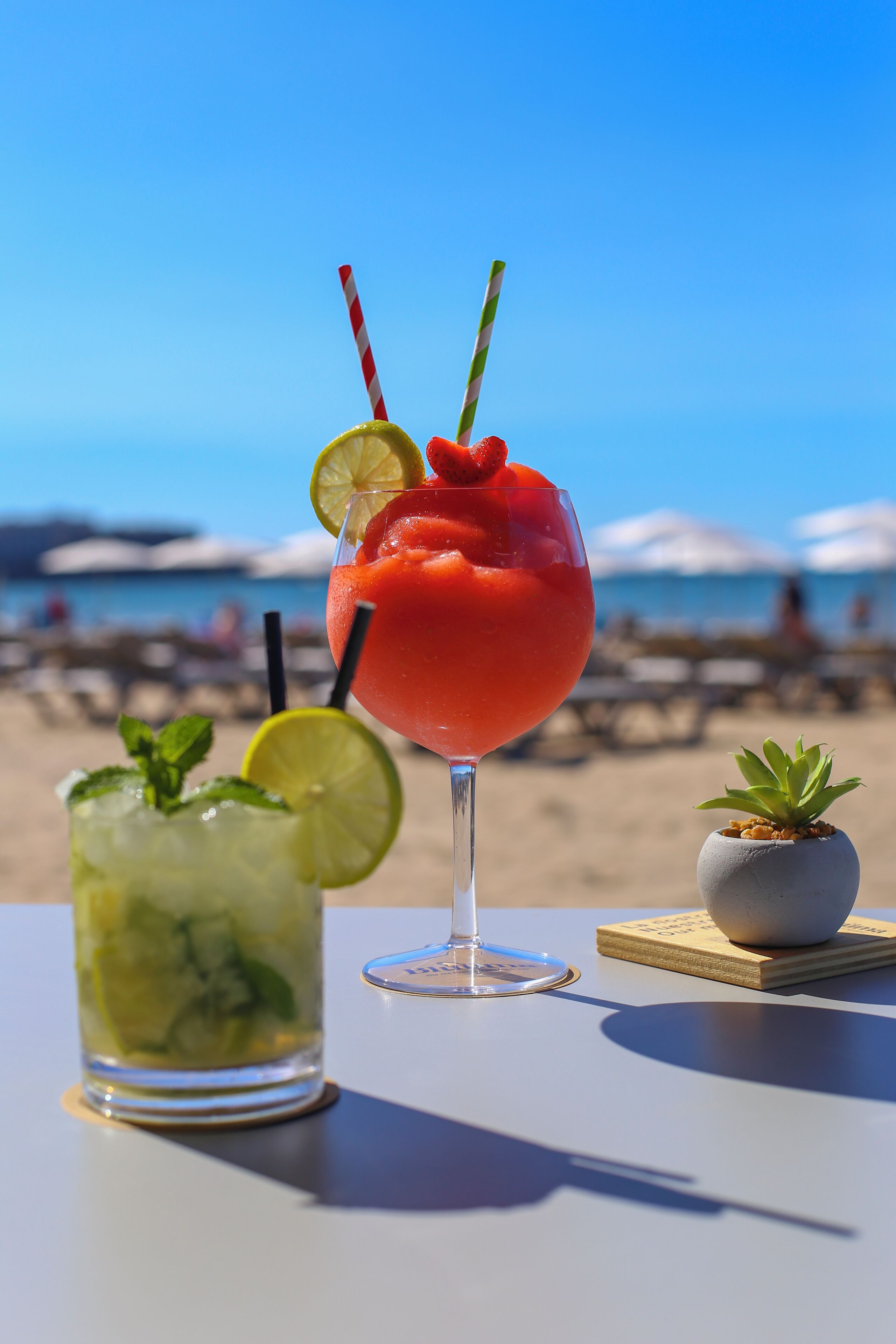 A table with a red cocktail, a green drink, and a small plant on a sunny beach.