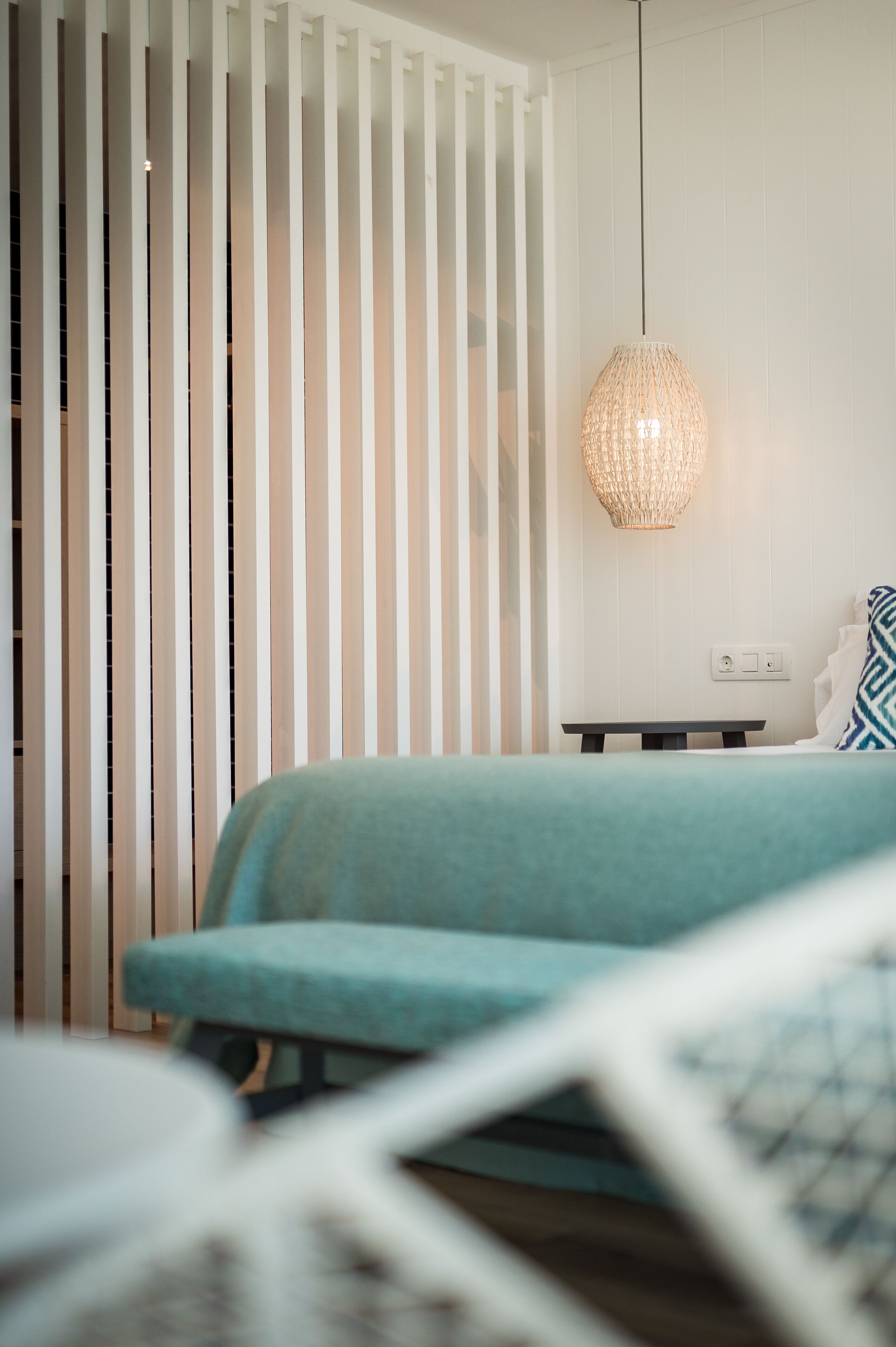 A bedroom with a bed, a bedside table, a hanging light fixture, and a white wall with vertical blinds.