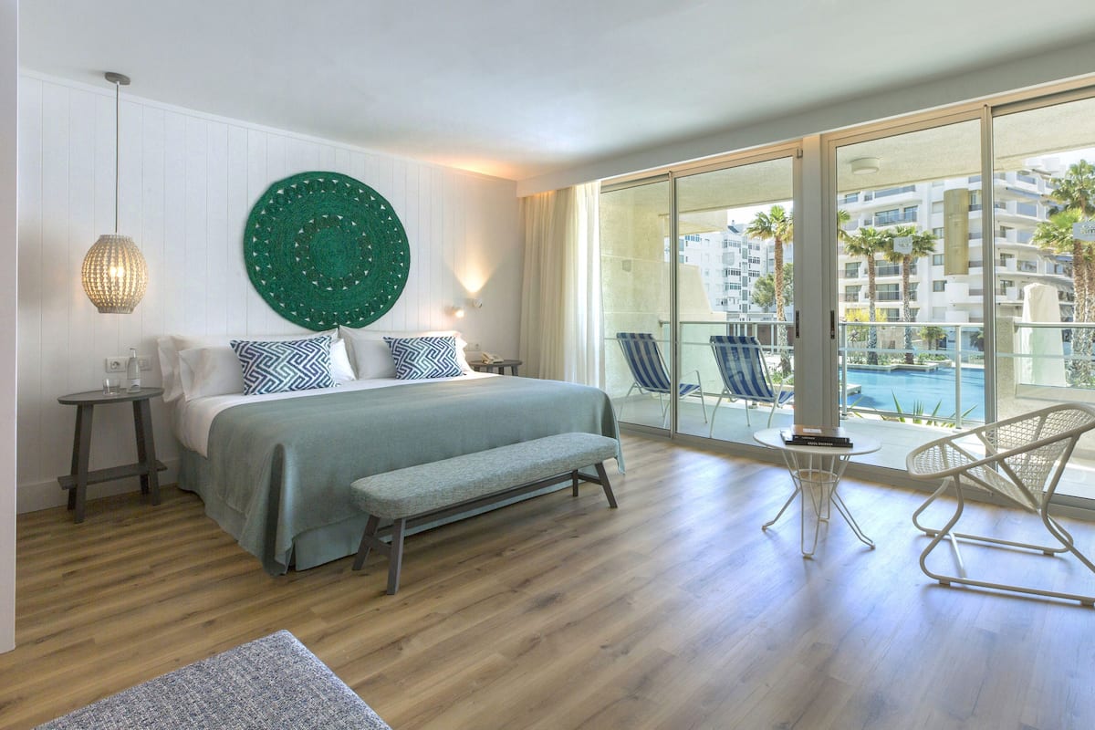 A hotel room with a large bed, a view of a swimming pool, and a balcony with chairs.