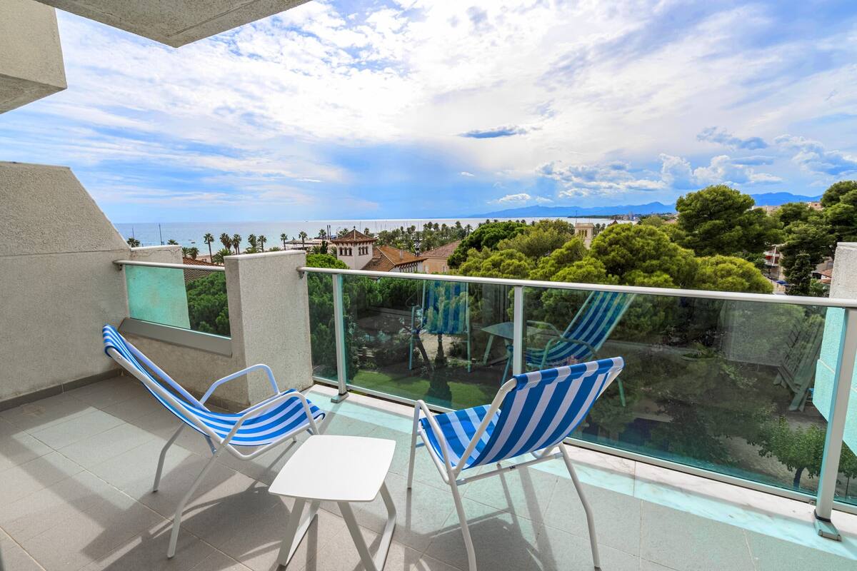 A balcony with two striped chairs and a small table, overlooking a pool and greenery.