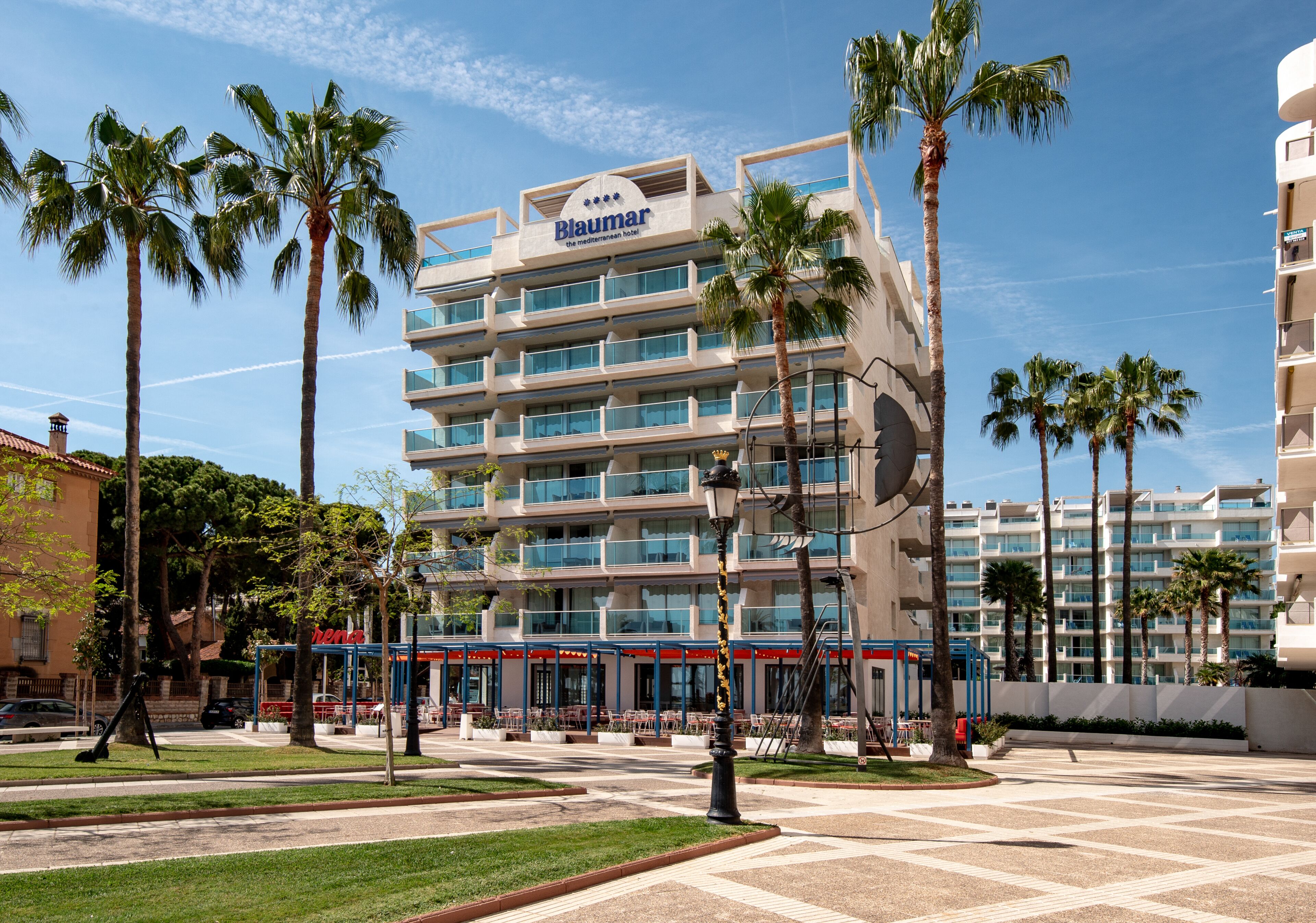 Un hotel de varios pisos con un letrero llamativo, palmeras y un patio empedrado.