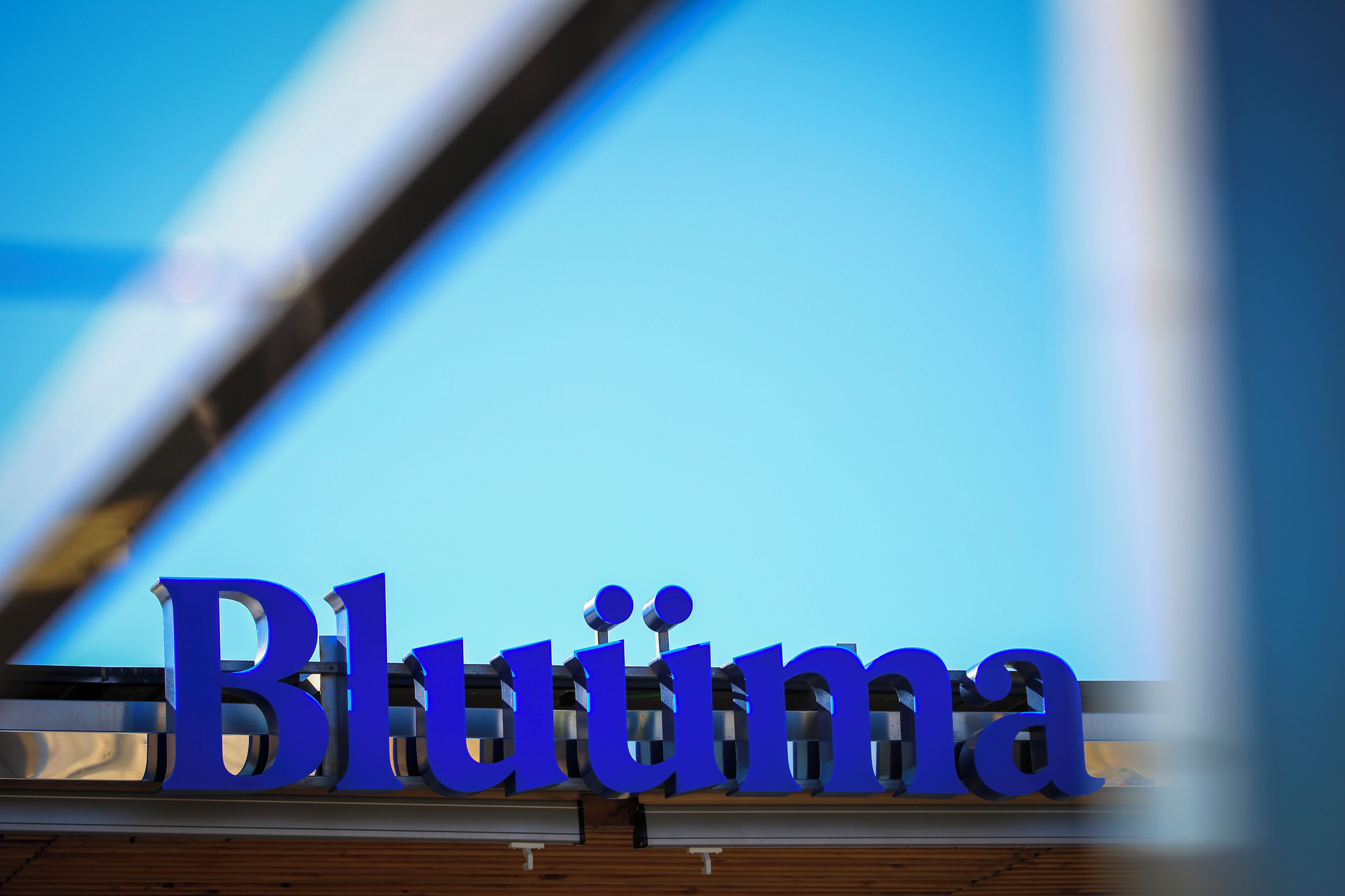 Een bord met de tekst 'Bluima' in grote blauwe letters, bevestigd aan een constructie met een houten basis.