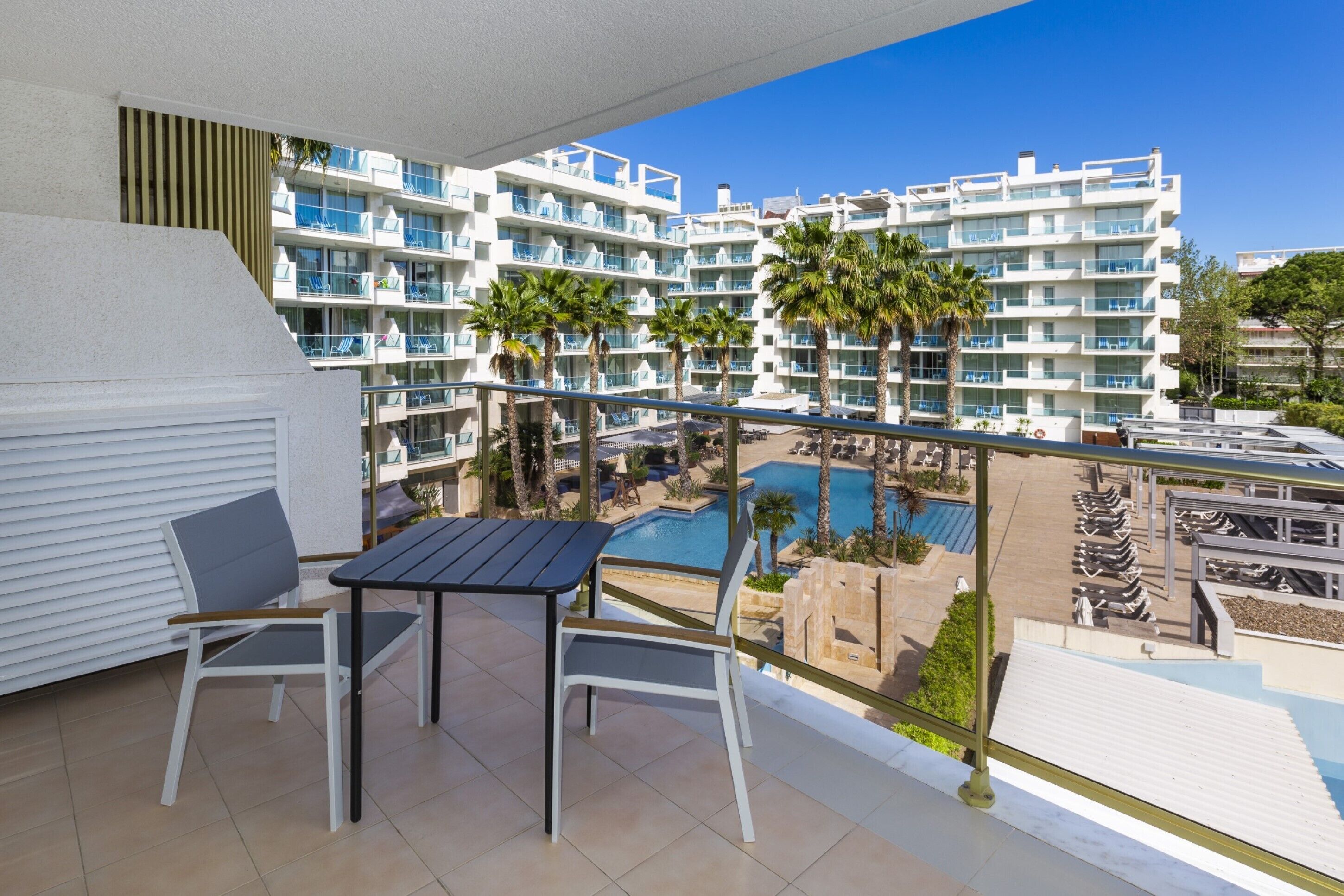 Un balcón con mesa y sillas, con vistas a una piscina y un edificio de resort con múltiples balcones y palmeras.