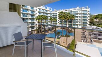Un balcón con mesa y sillas, con vistas a una piscina y un edificio de resort con múltiples balcones y palmeras.
