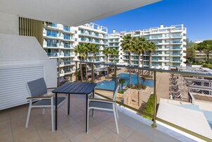 Un balcón con mesa y sillas, con vistas a una piscina y un edificio de resort con múltiples balcones y palmeras.