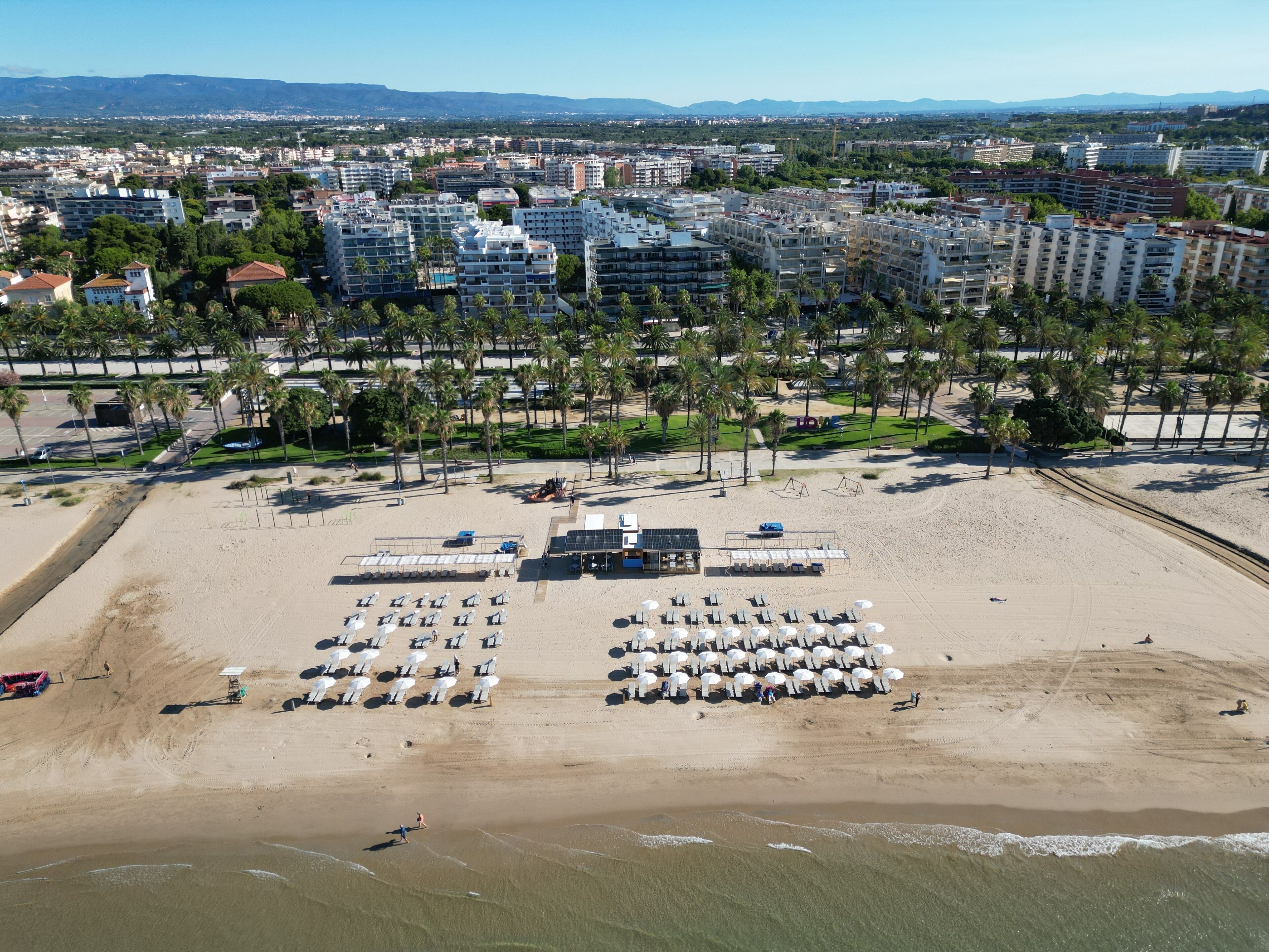 Eine Luftaufnahme eines Strandes mit Reihen von Liegestühlen und Sonnenschirmen, einer kleinen Bühne und einem Sandstrand.
