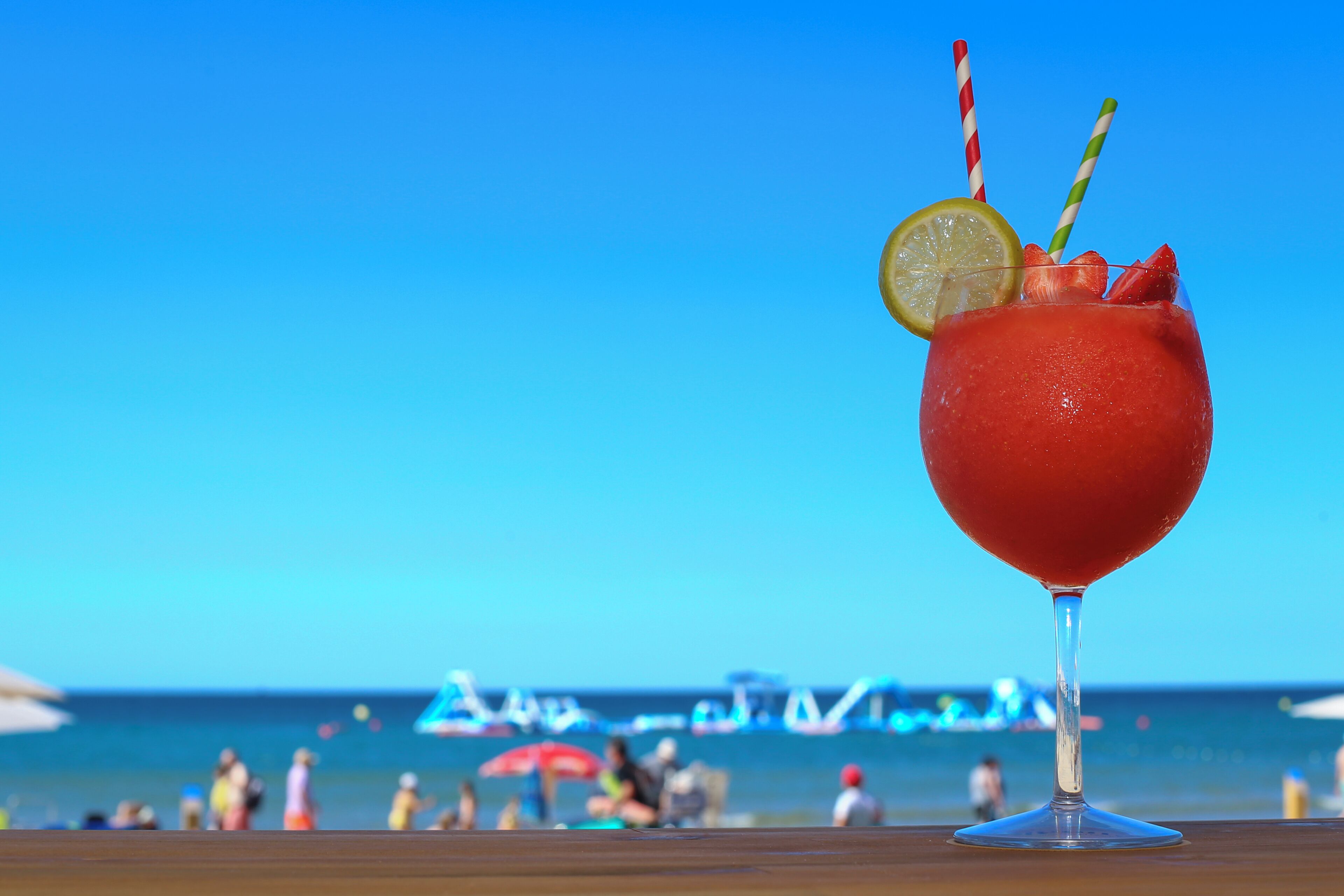 A cocktail glass with a red drink, lemon slice, and striped straws on a wooden table with a beach and ocean in the background.