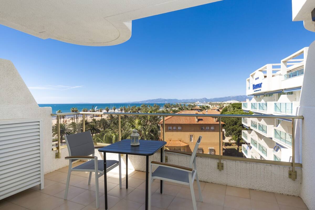 A balcony with a table and chairs, overlooking a beach and buildings.