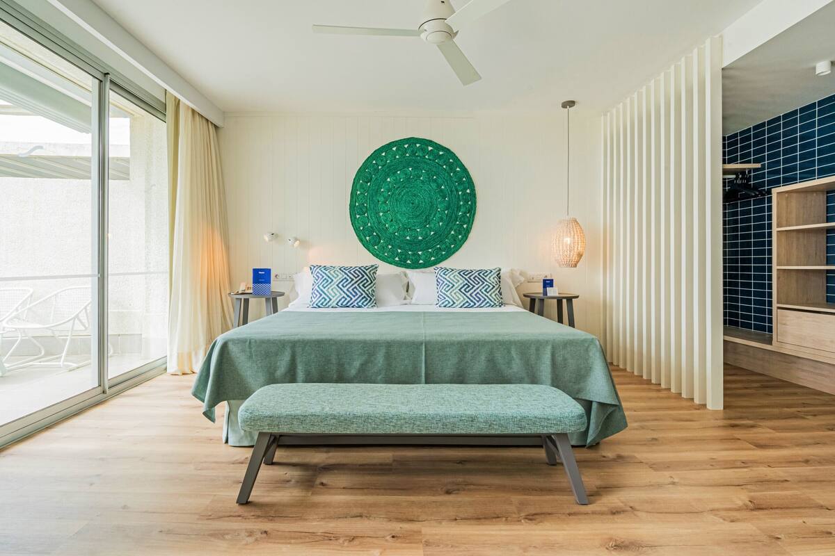 A modern bedroom with a large bed, a ceiling fan, a green wall hanging, and a view of the outdoors.