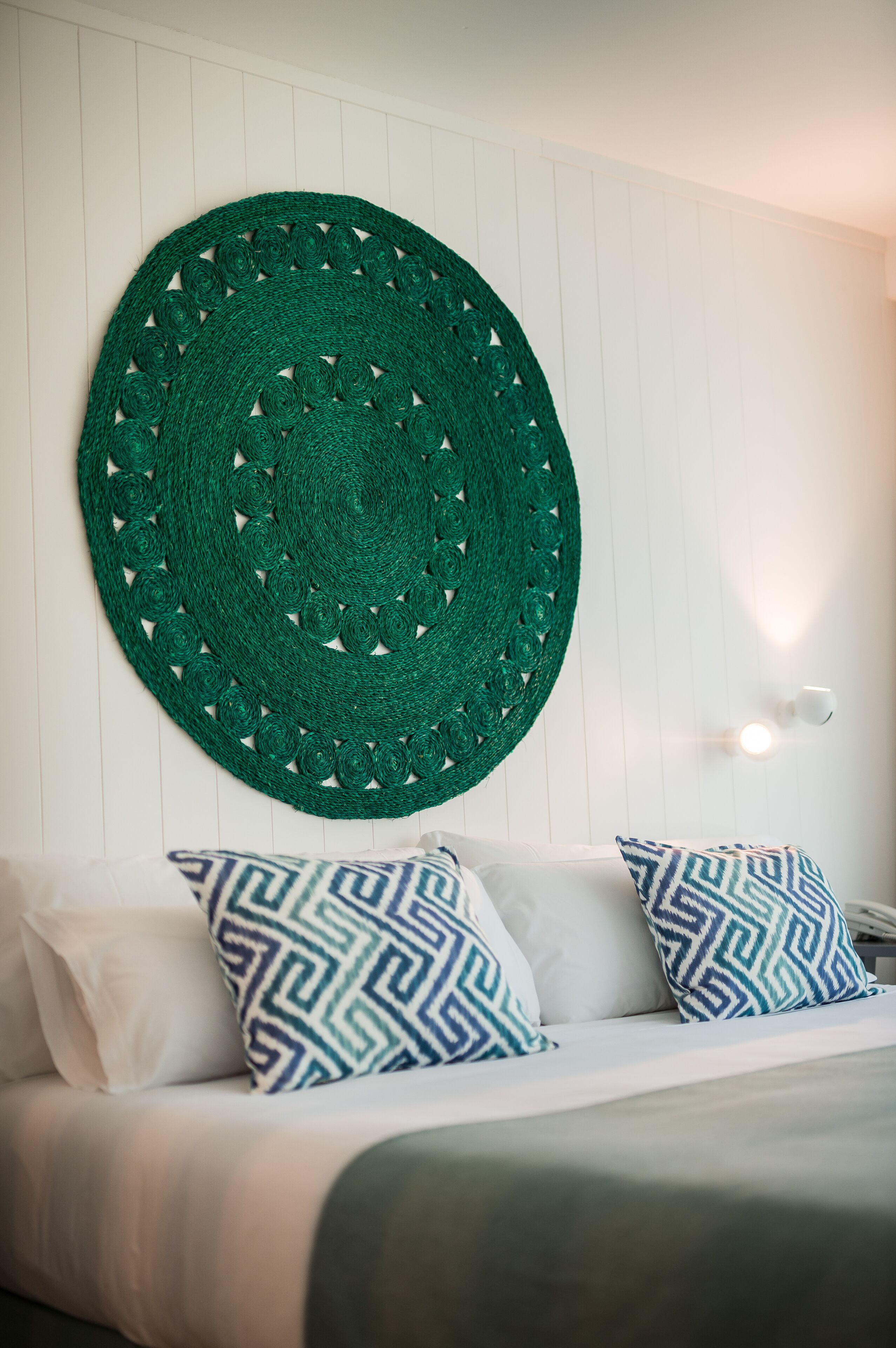 Un lit avec une literie blanche et des coussins à motifs, une décoration murale tissée de couleur verte et un mur blanc agrémenté d’un luminaire.