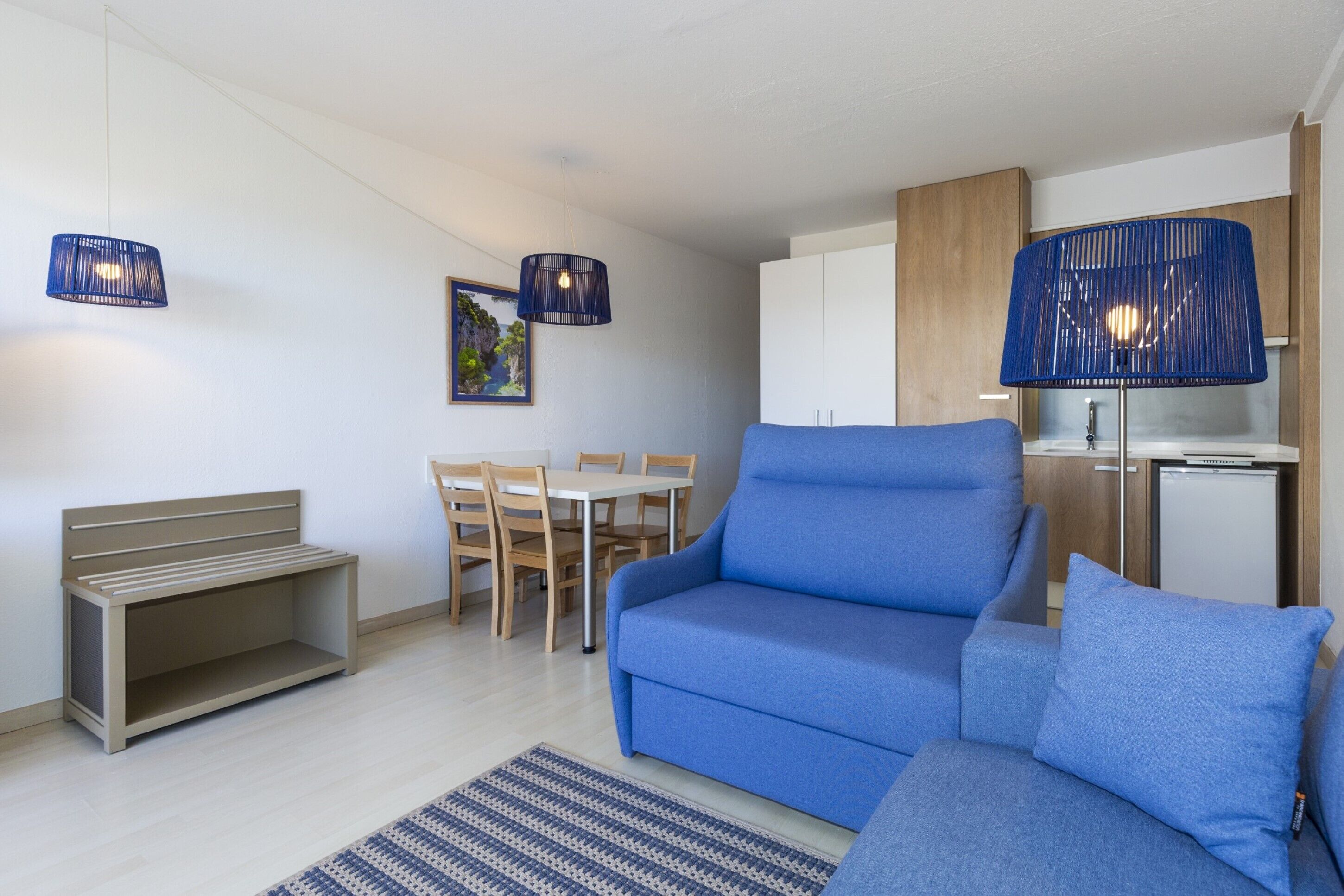 A modern living room with a blue sofa, a wooden bench, a dining table with chairs, and a kitchen area with cabinets and a refrigerator.