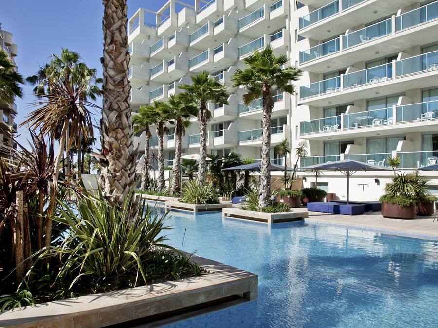 Un hotel moderno con zona de piscina, palmeras y mobiliario exterior.