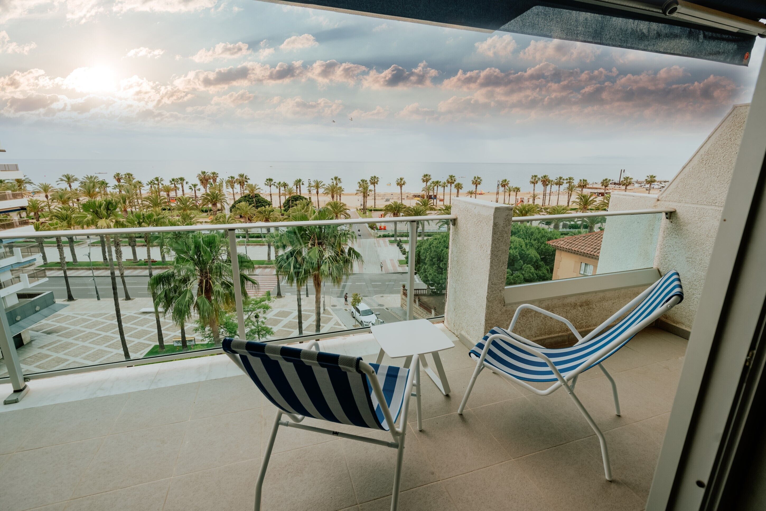 A balcony with two striped chairs and a small table, overlooking a beach with palm trees and a clear sky.