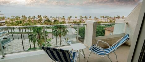 A balcony with two striped chairs and a small table, overlooking a beach with palm trees and a clear sky.