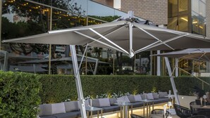 Un área de estar al aire libre con mesas y sillas, un paraguas grande, y un edificio con un letrero que dice 'HOTEL AMBASSADOR'.