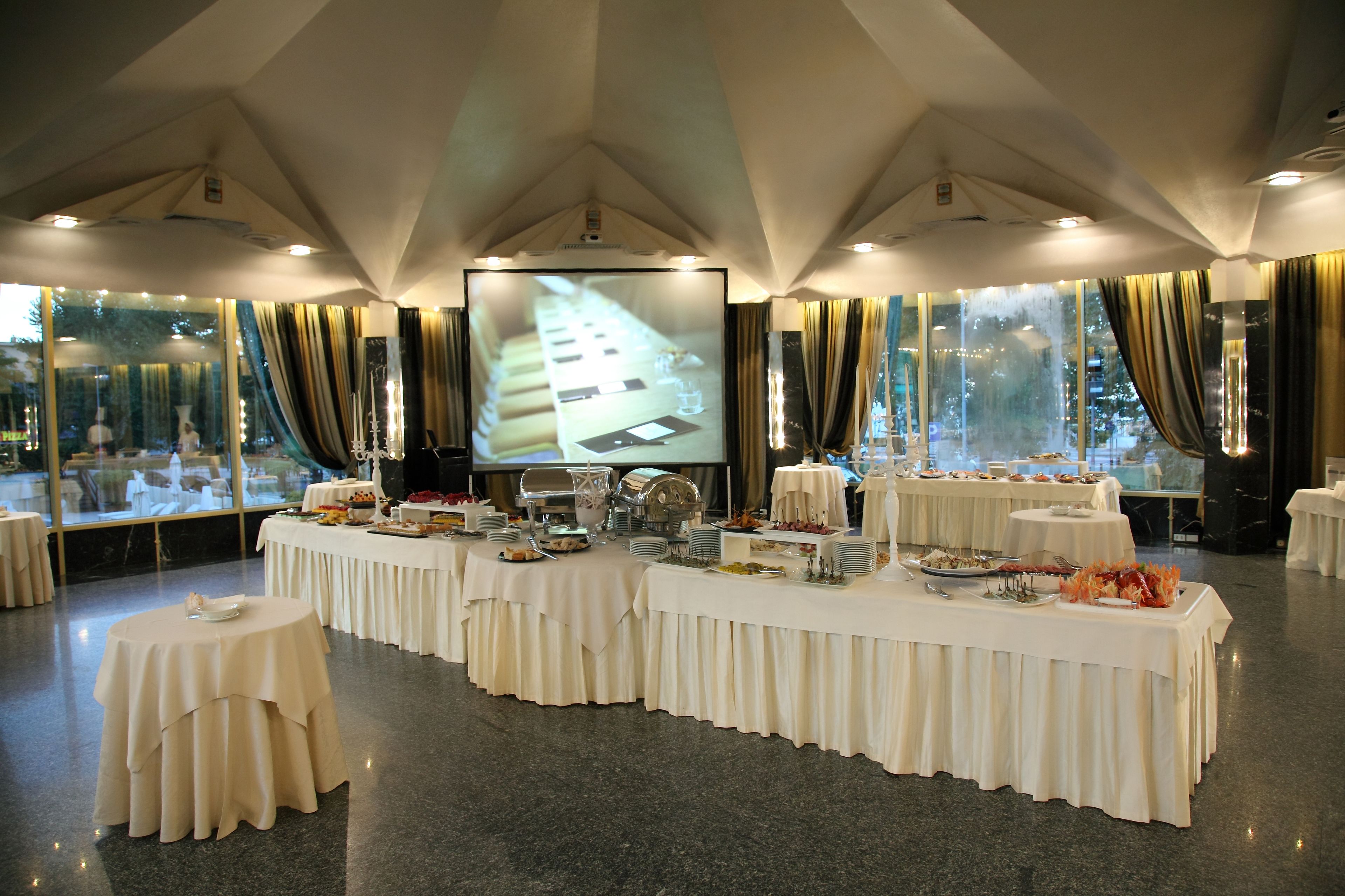 Un salón de banquetes con mesas dispuestas para una comida, una pantalla grande mostrando una presentación y amplias ventanas con cortinas.