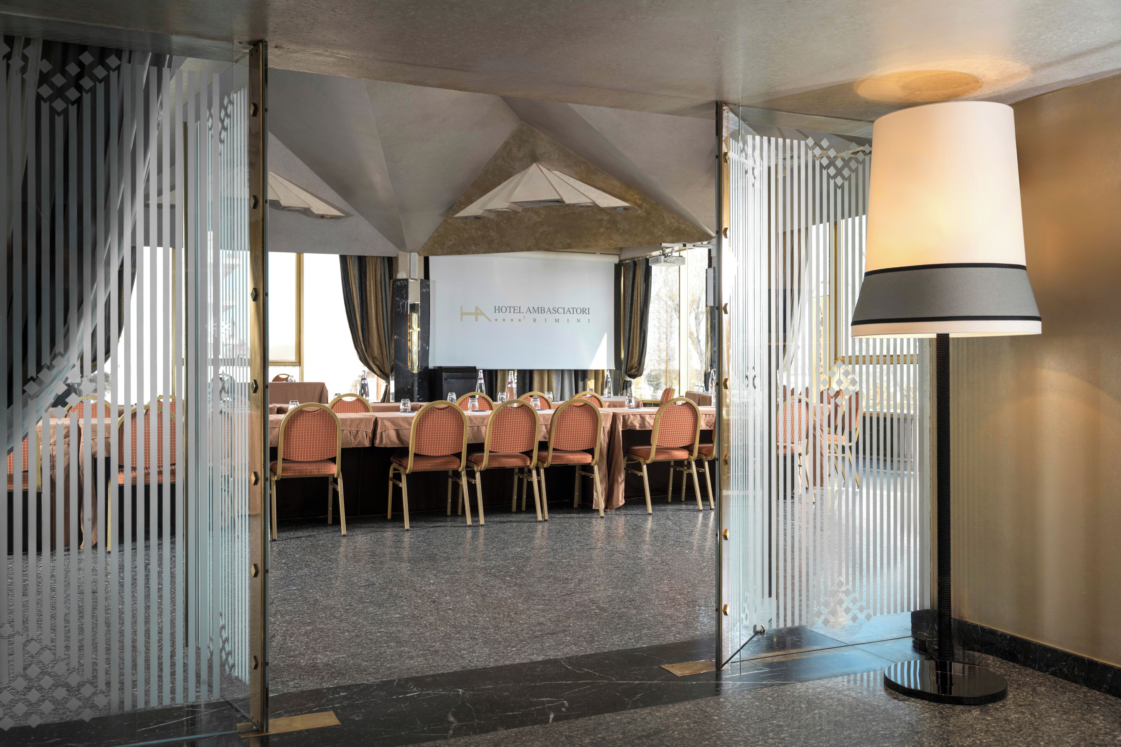A conference room with a long table, chairs, a projector screen, and a floor lamp.